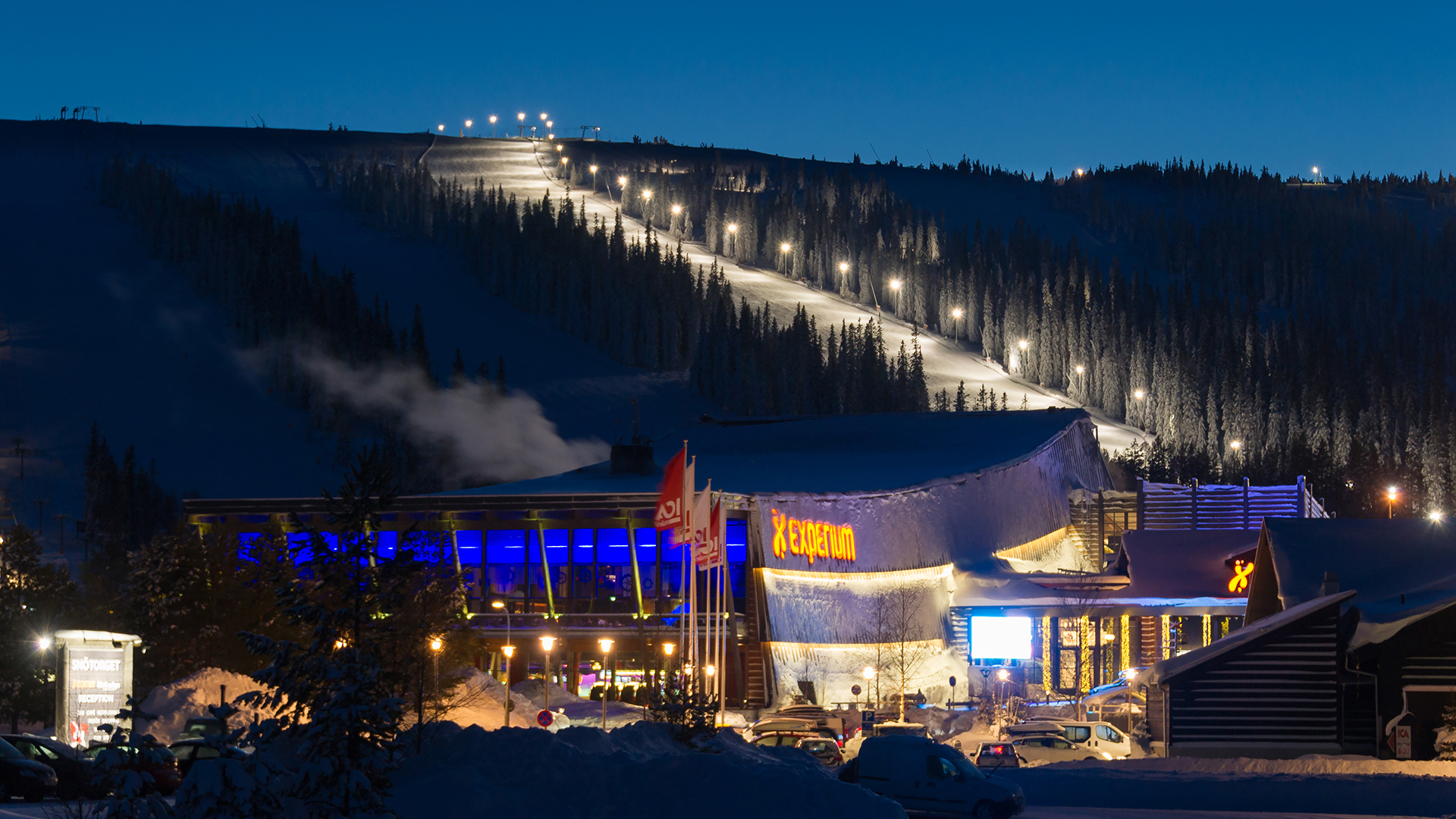 Kvällsvy över ett upplyst skidcenter i fjällmiljö, med snöbeklädda pister och modernt servicehus