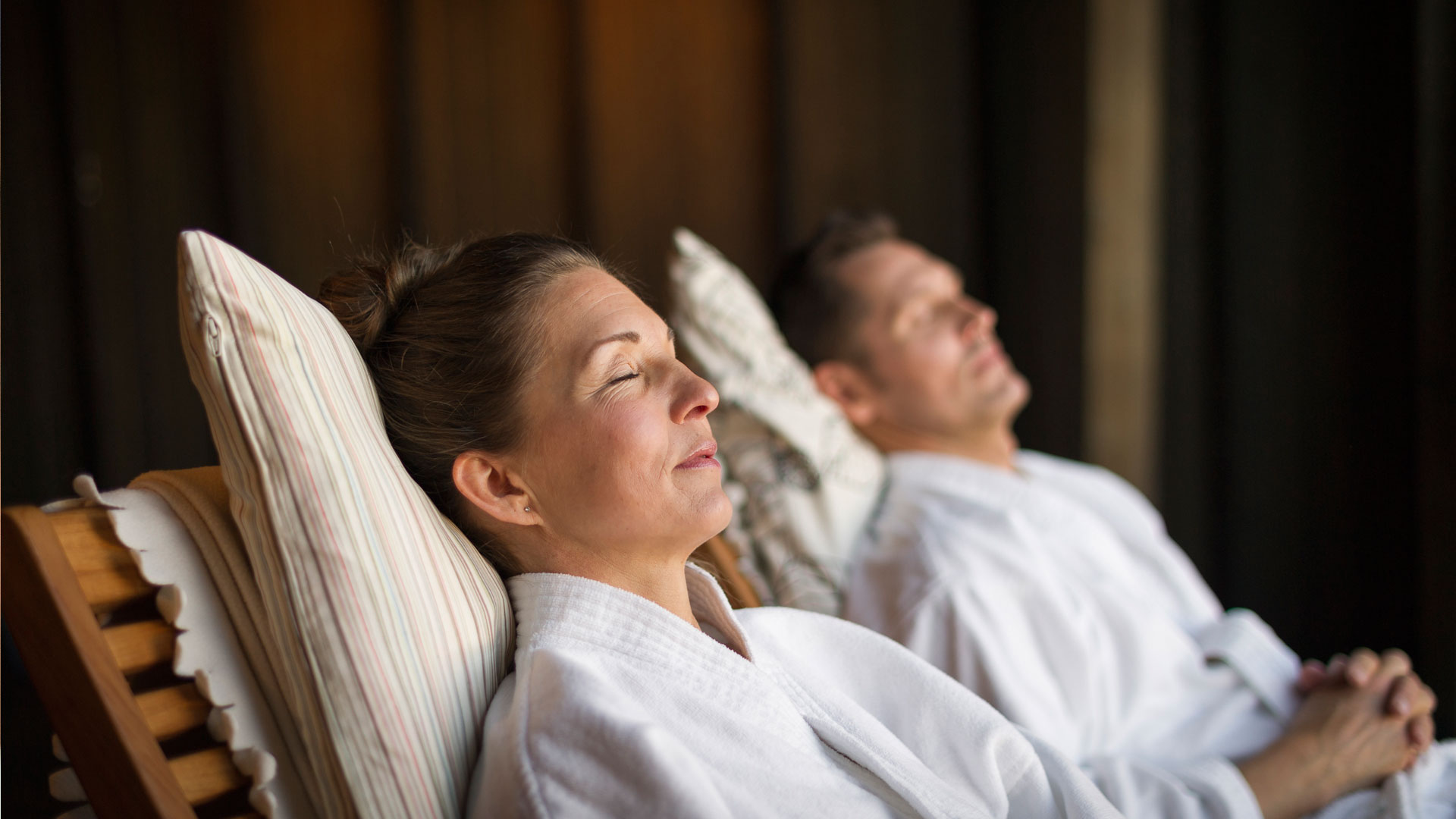 Två personer slappnar av i en spa-miljö, iklädda vita badrockar, liggandes på solstolar med stängda ögon