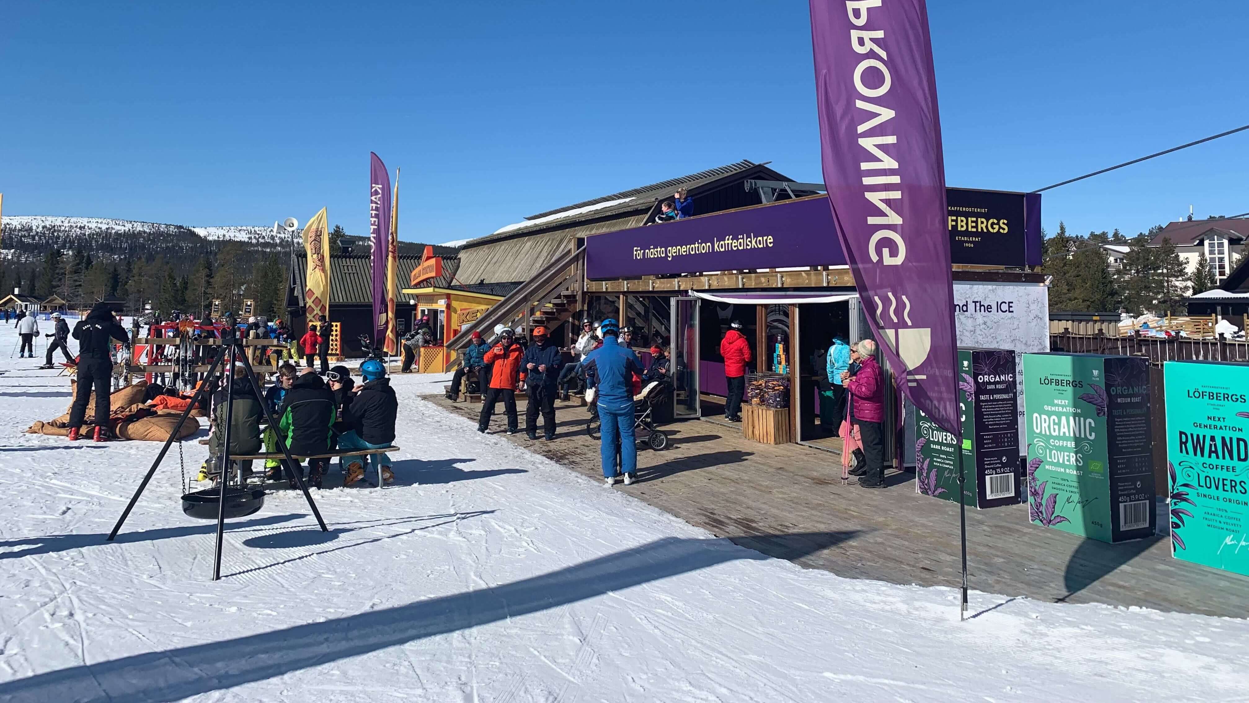 Skiers gather at a coffee cabin in the sunshine at a ski resort, with snow-covered mountains in the background and colorful flags adorning the area