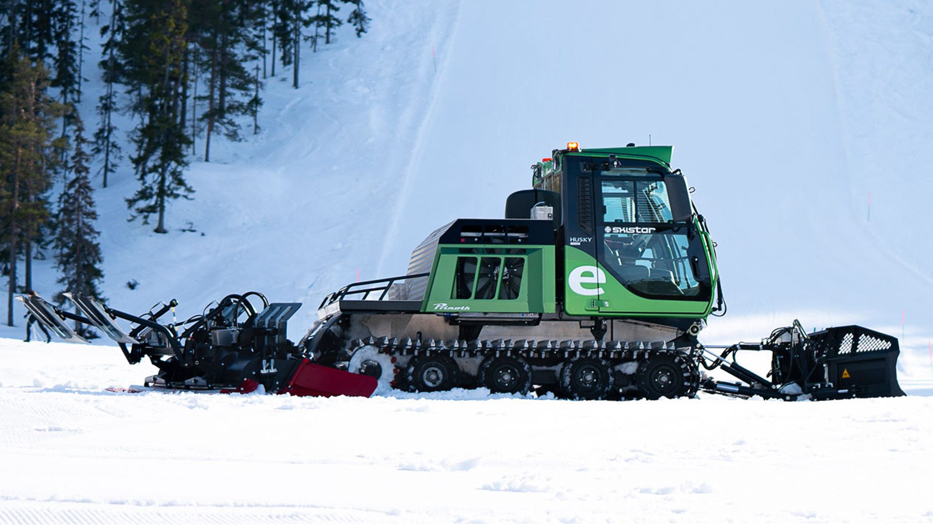Grön pistmaskin som preparerar snö på en skidbacke, omgiven av snötäckta träd och ljusblå himmel