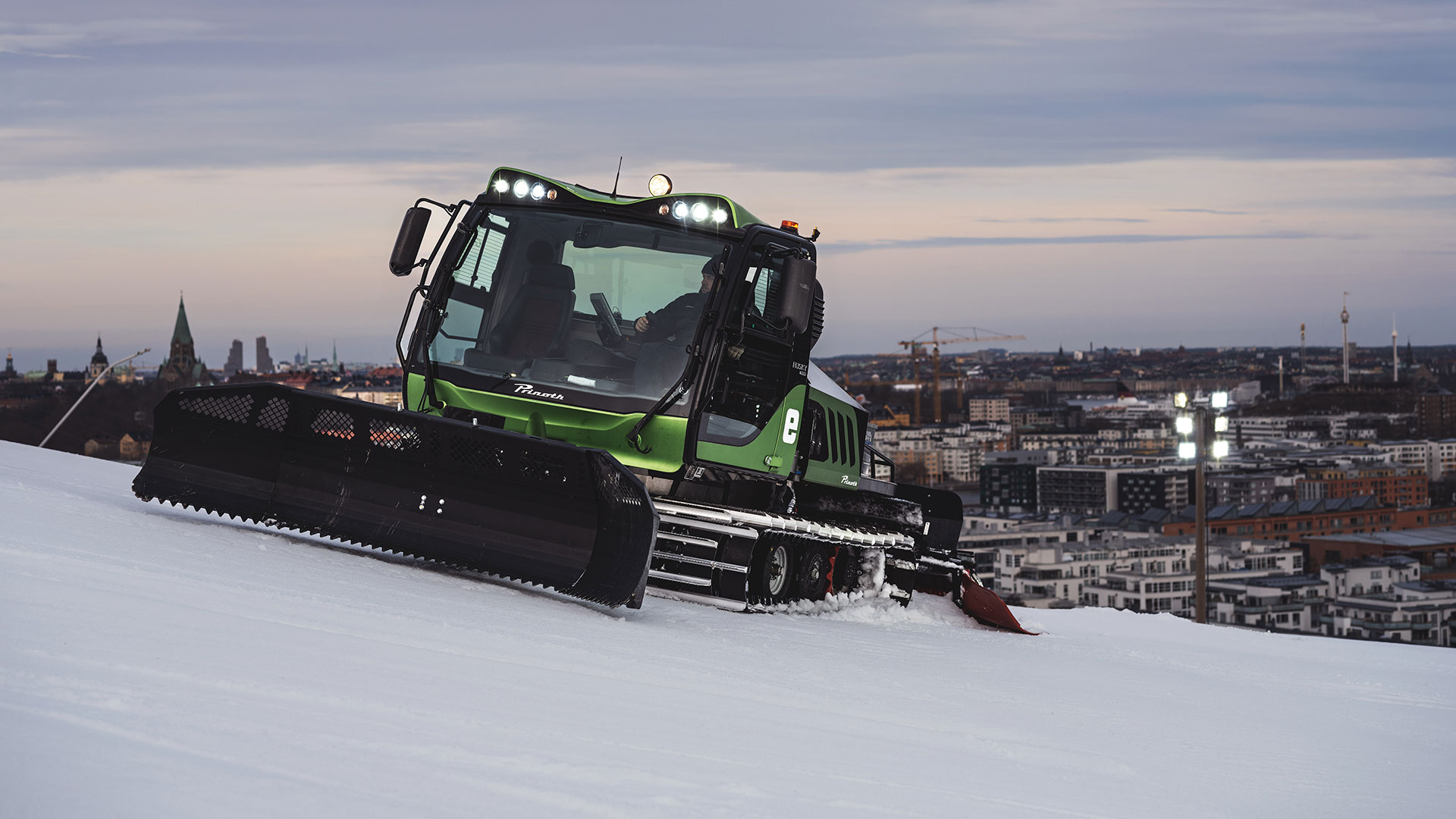 Snöplogmaskin i arbete på en snötäckt backe med stadsutsikt i bakgrunden