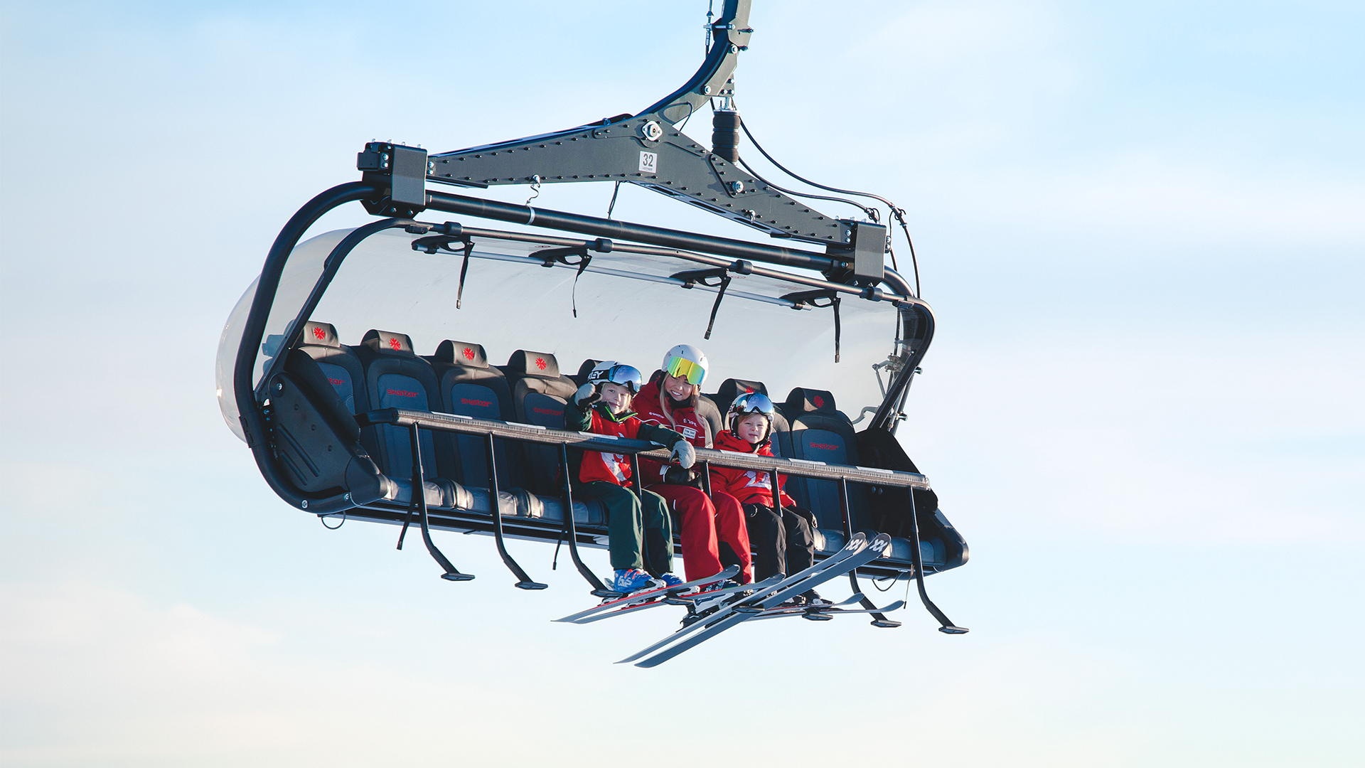 Personer på en sittlift i skidbacken med klarblå himmel i bakgrunden. De bär skidkläder och hjälmar för säkerhet på skidresan