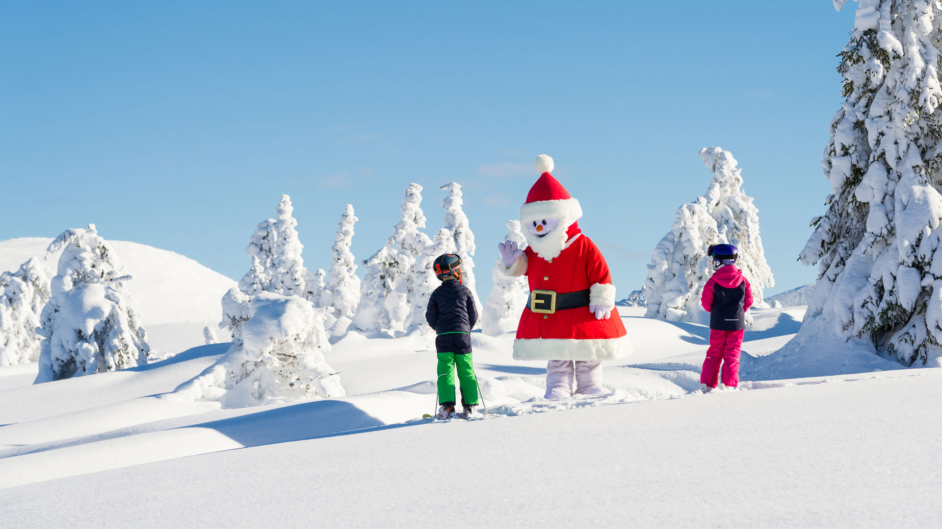 Kinder beim Skifahren mit Schneemann Valle in Weihnachtsmann-Kostüm in verschneiter Landschaft – ein Bild von Weihnachten in den Bergen.