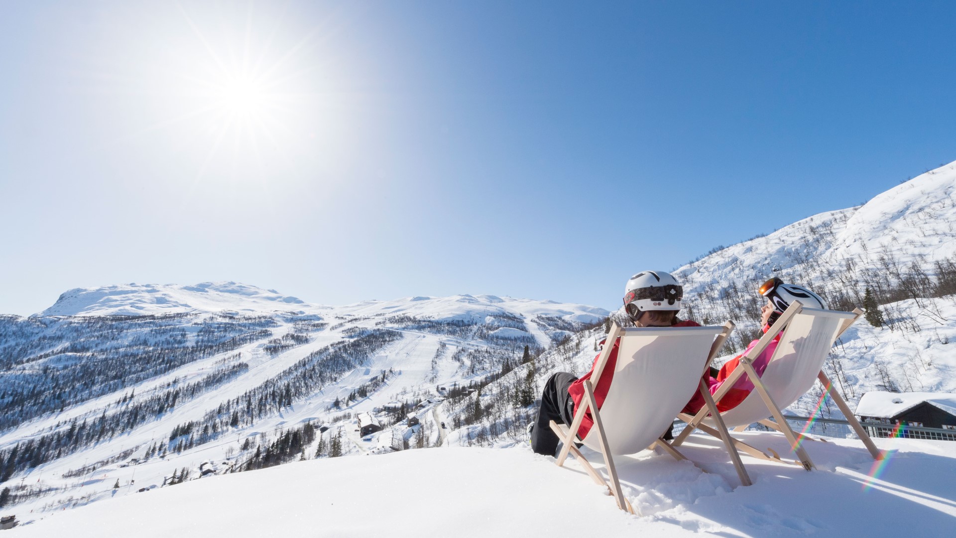 Två personer sitter i solstolar på en snötäckt fjälltopp i fjällen, med hjälmar och skidkläder, njuter av solskenet och vacker utsikt under påsklovet.