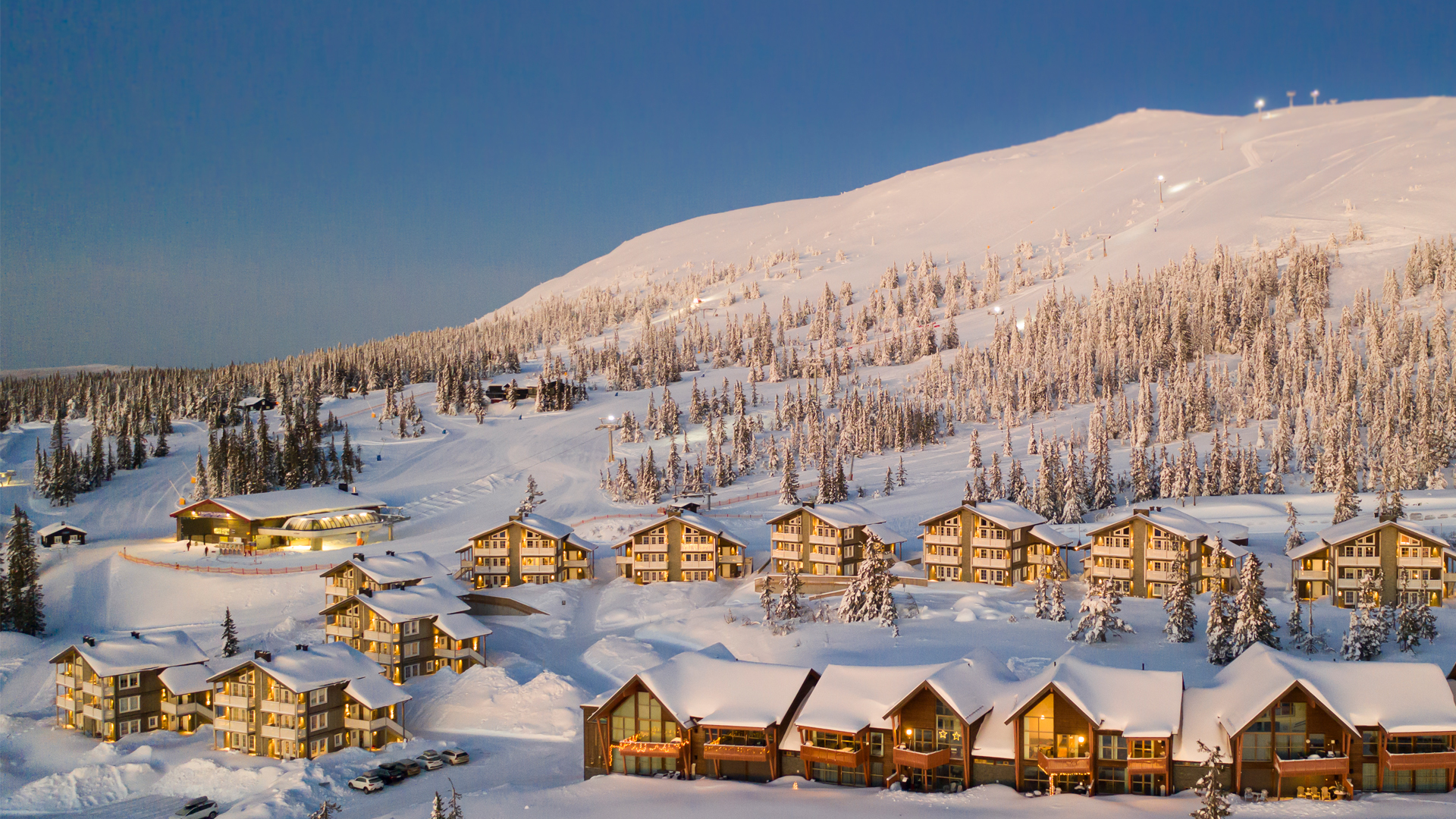 Vinterlandskap med snötäckta stugor och skidbackar, omgivna av snöklädda träd, under en klar himmel i fjällmiljö