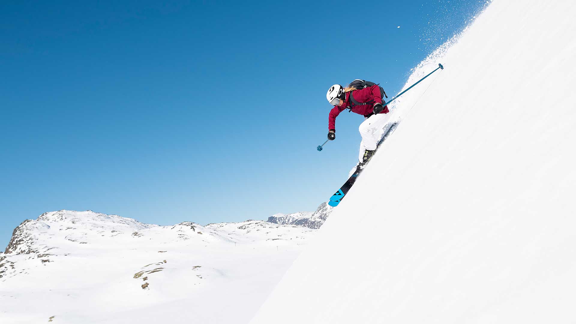 Person på ski ned en bratt snødekt fjellside under en blå himmel