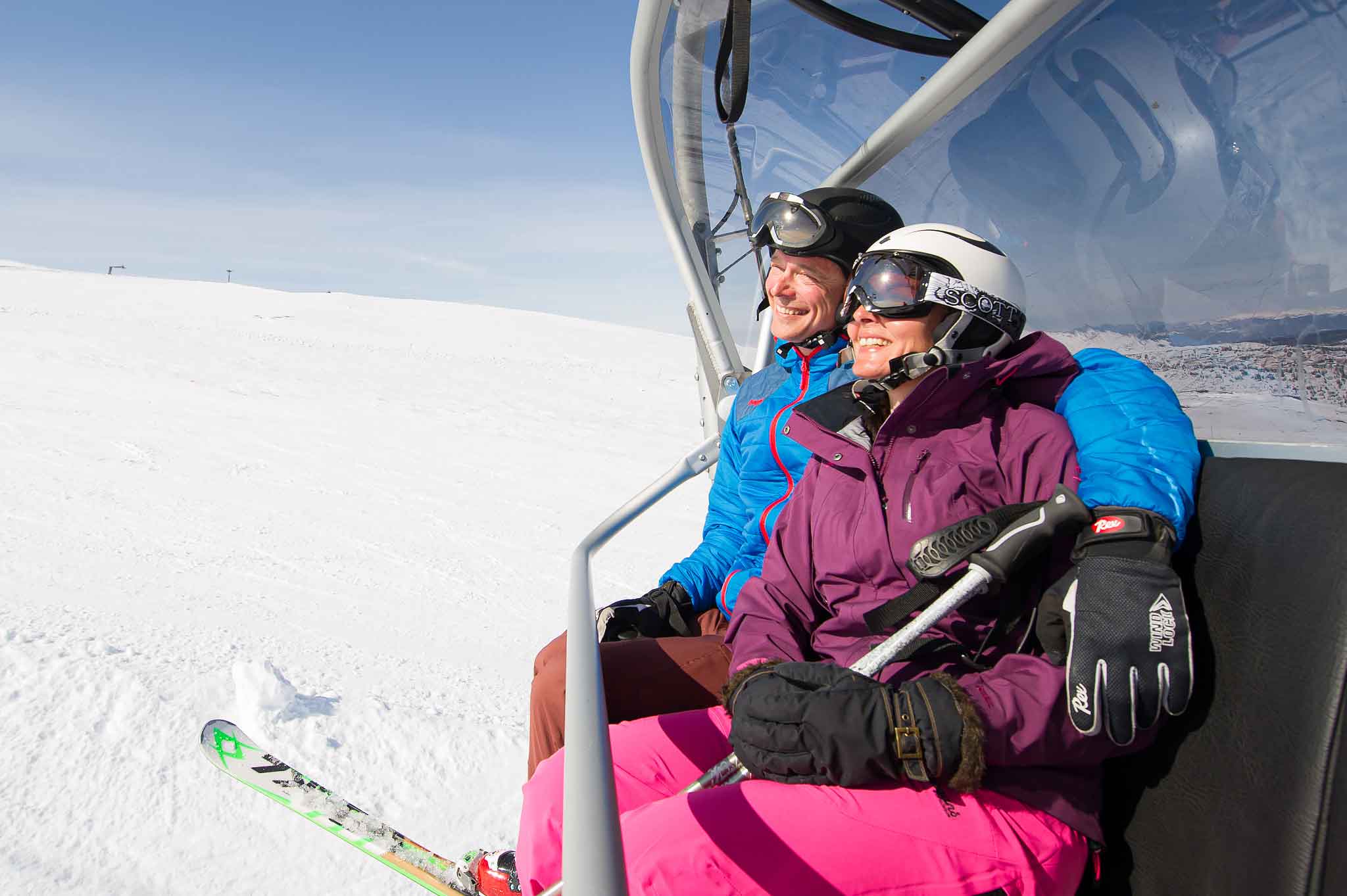 To personer i fargerike skiklær sitter sammen i en skiheis på en solfylt dag i fjellene. Snøen er skinnende hvit under dem, og de ser fornøyde ut mens de nyter turen