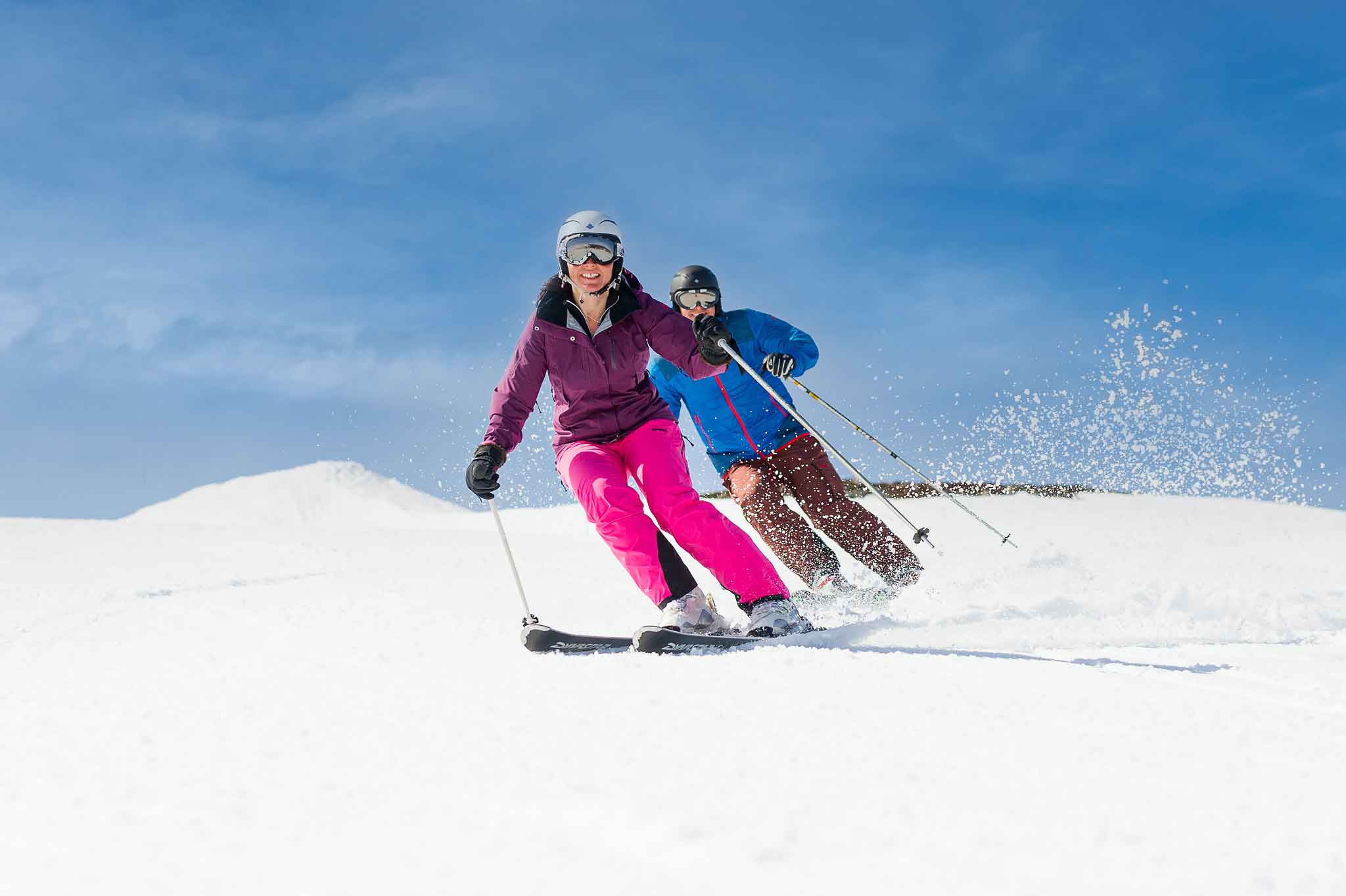 To personer på ski i bakken under en klar blå himmel, iført vinterklær og skiutstyr