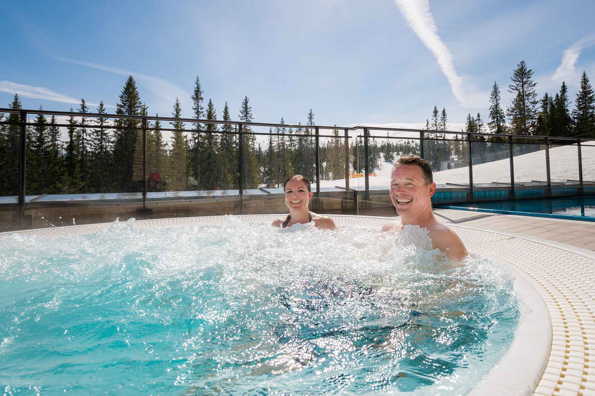 Par i boblebad med utsikt mot snødekte trær og klar blå himmel, nyter solfylt vinterdag i norsk fjellområde