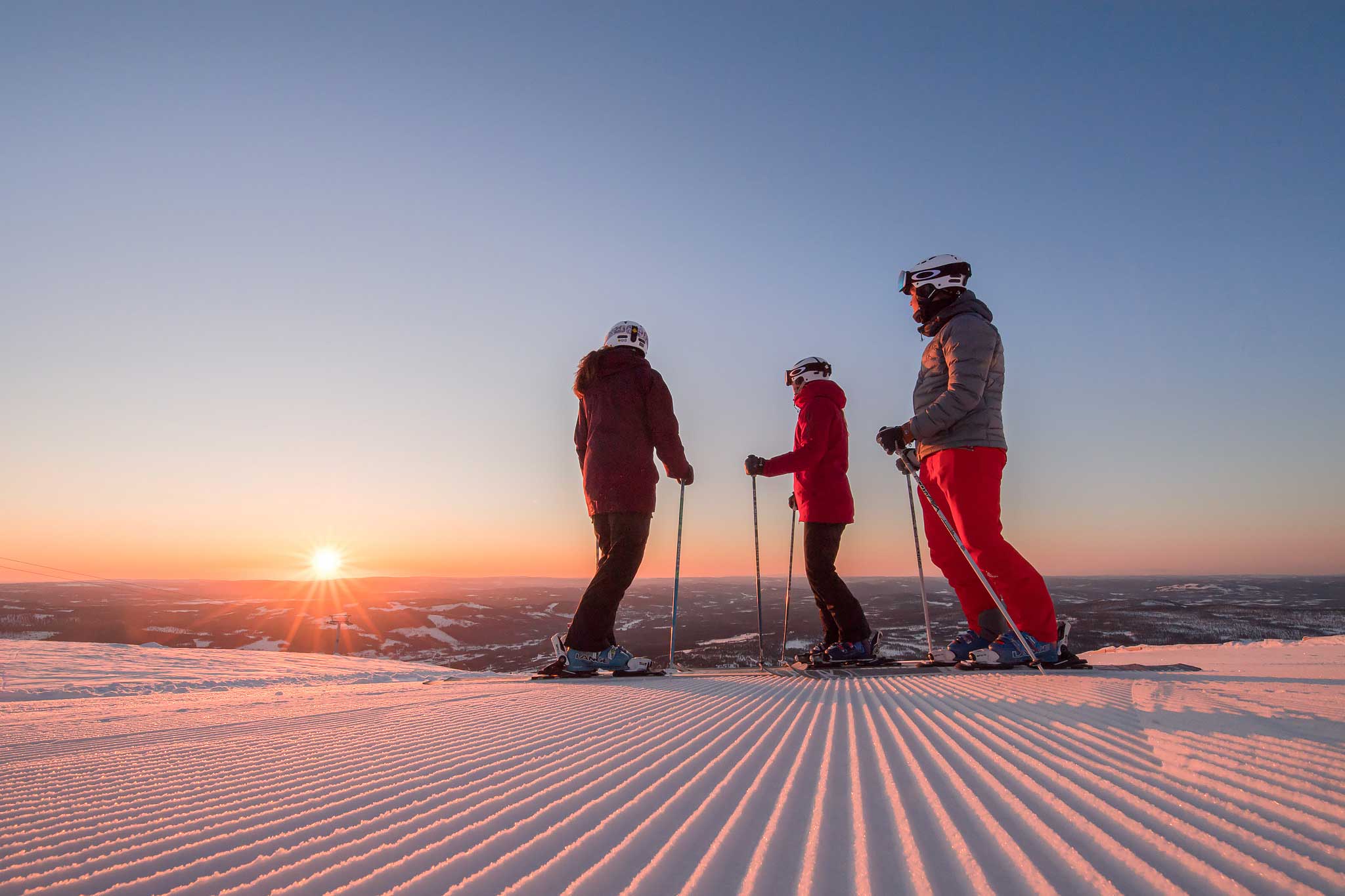 Tre personer på ski nyter solnedgangen på en snødekt fjelltopp, iført skiutstyr og hjelmer