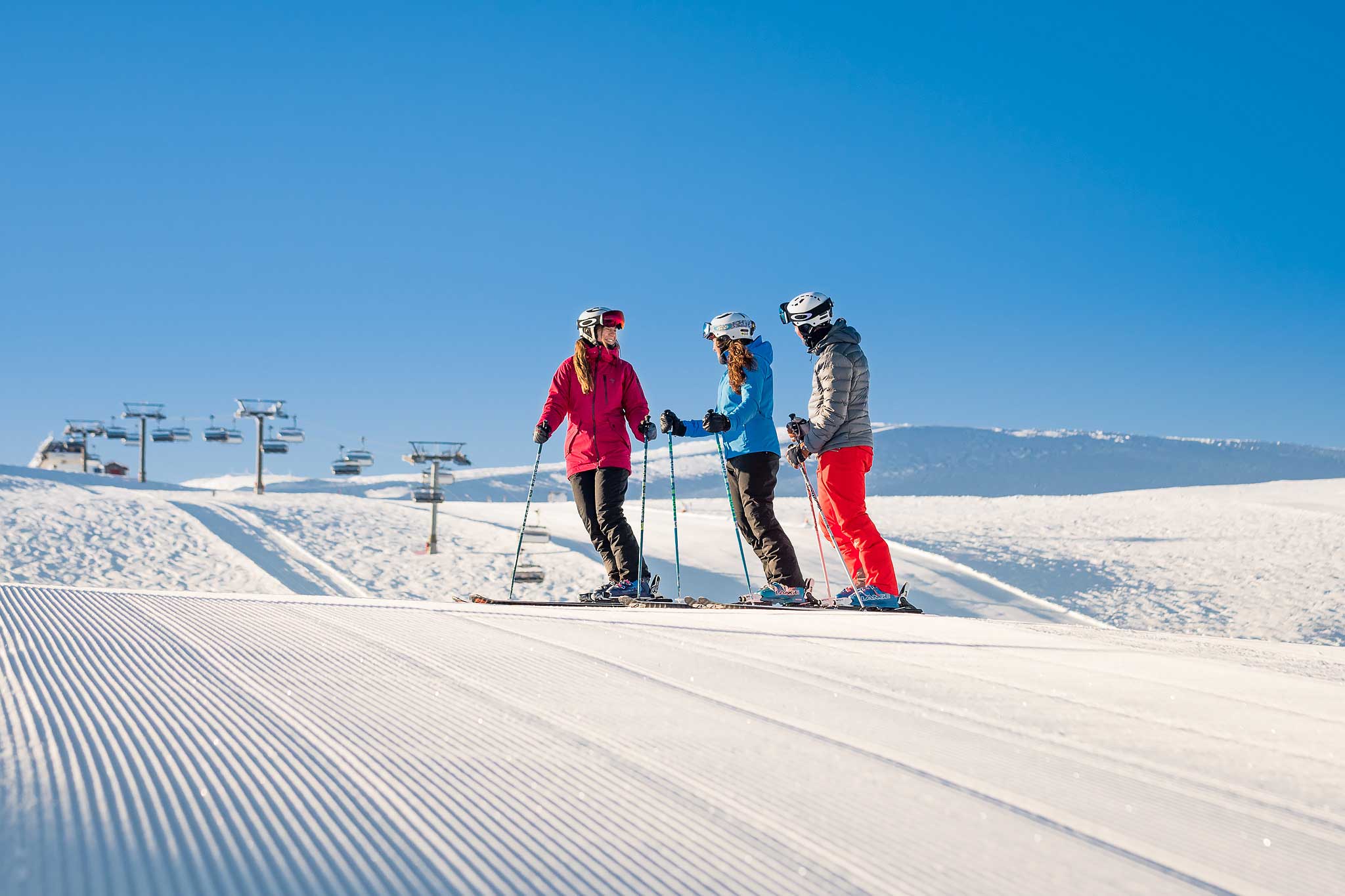 Tre personer står på ski i en preparert bakke under en klar blå himmel, med skiheiser i bakgrunnen