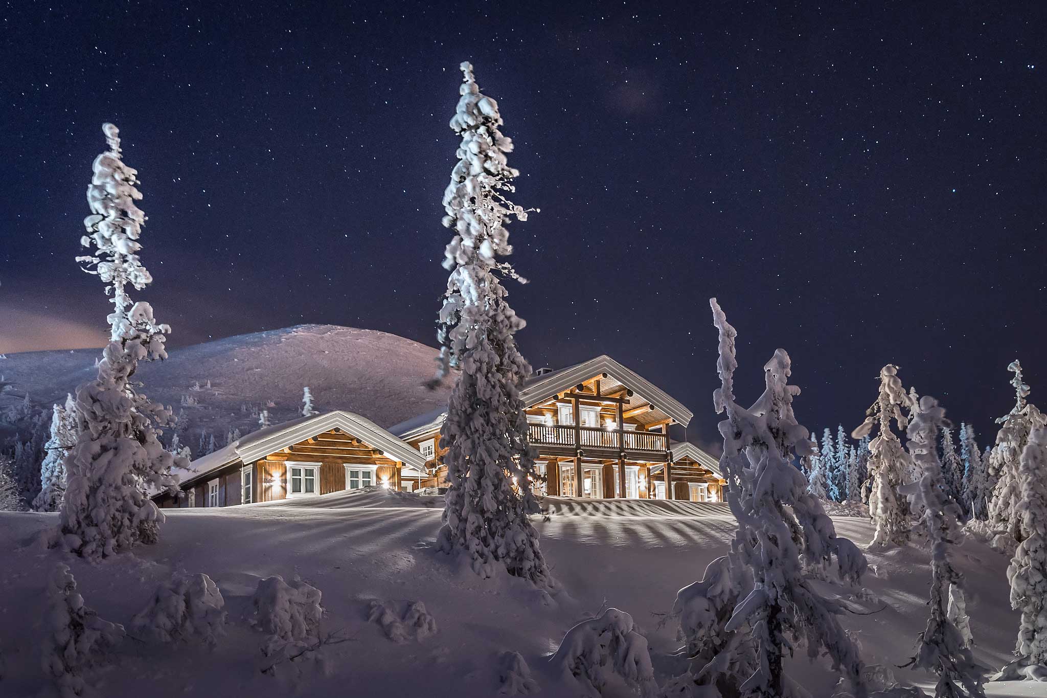 Snødekt trehytte i naturskjønne omgivelser om natten, omringet av stjerner og snødekte trær