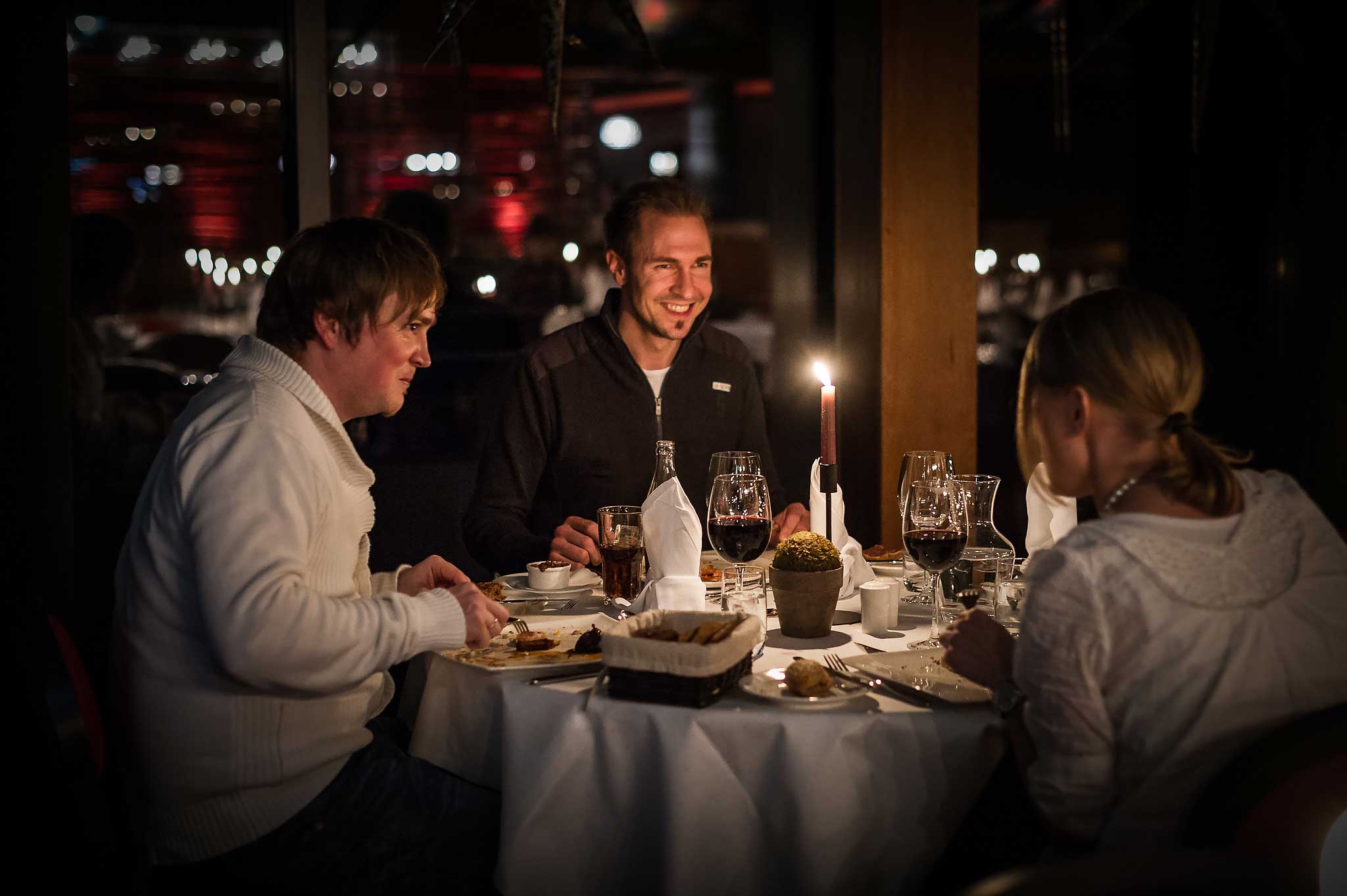 Tre personer sitter rundt et bord på en koselig restaurant. De nyter middag med stearinlys, vin og elegant borddekking. Atmosfæren er varm og avslappet