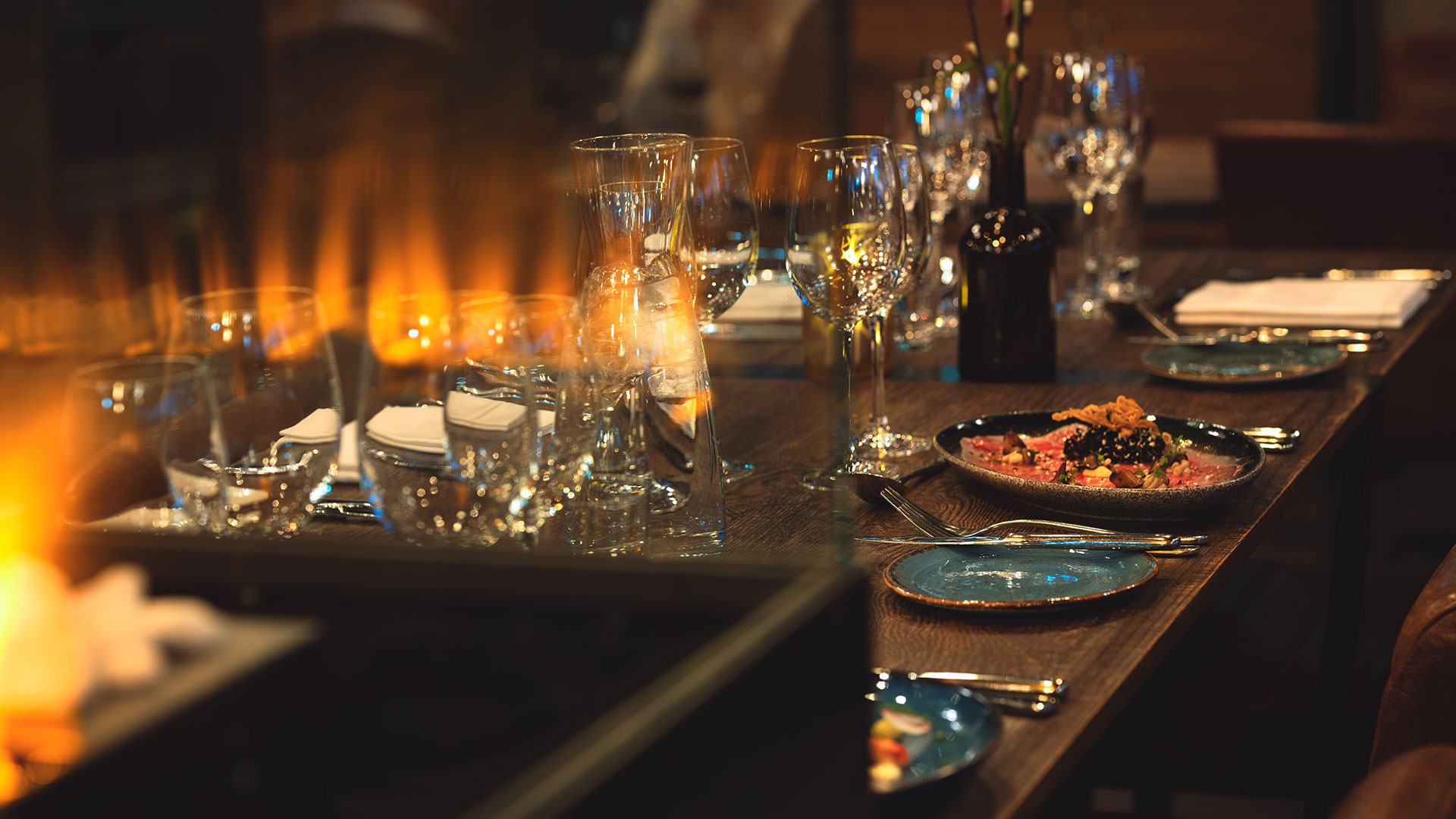 Elegant dukning på en restaurang med glas, tallrikar och en dekorerad maträtt, i bakgrunden syns en brinnande eld vilket skapar en varm och inbjudande atmosfär