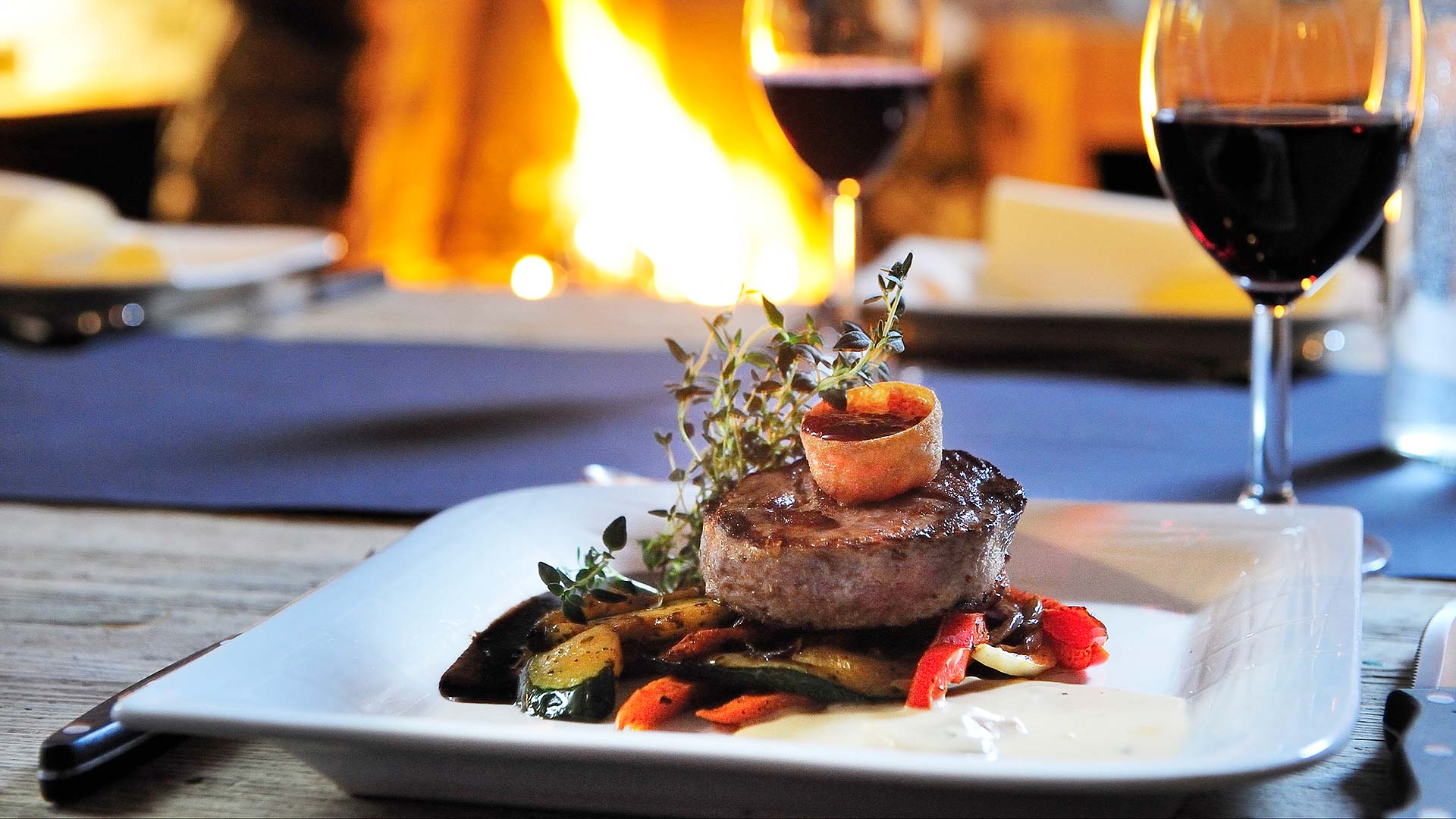 Biffrett med grønnsaker, saus og urter på en hvit tallerken, akkompagnert av et glass rødvin, foran en varm peis