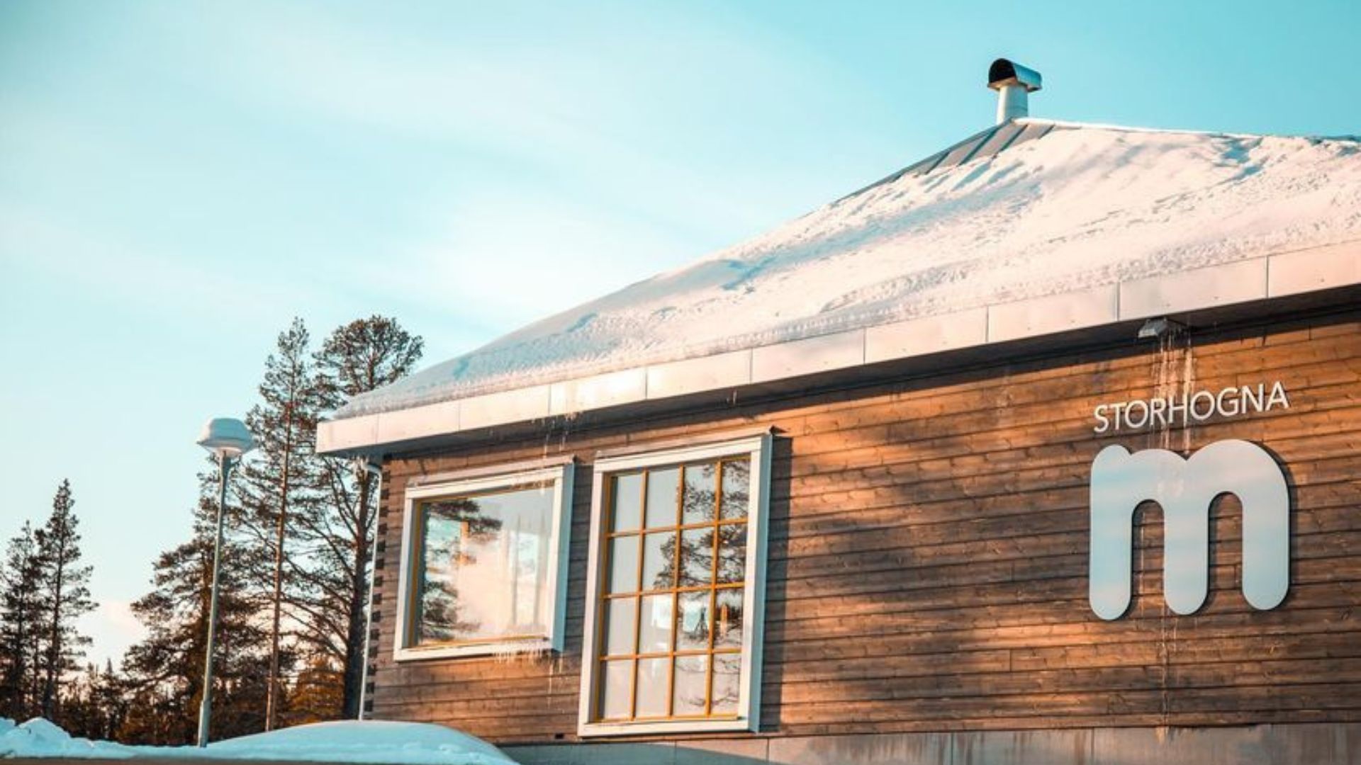 Byggnad med snötäckt tak och texten "Storhogna" på fasaden, omgiven av snötäckta granar under en klar himmel