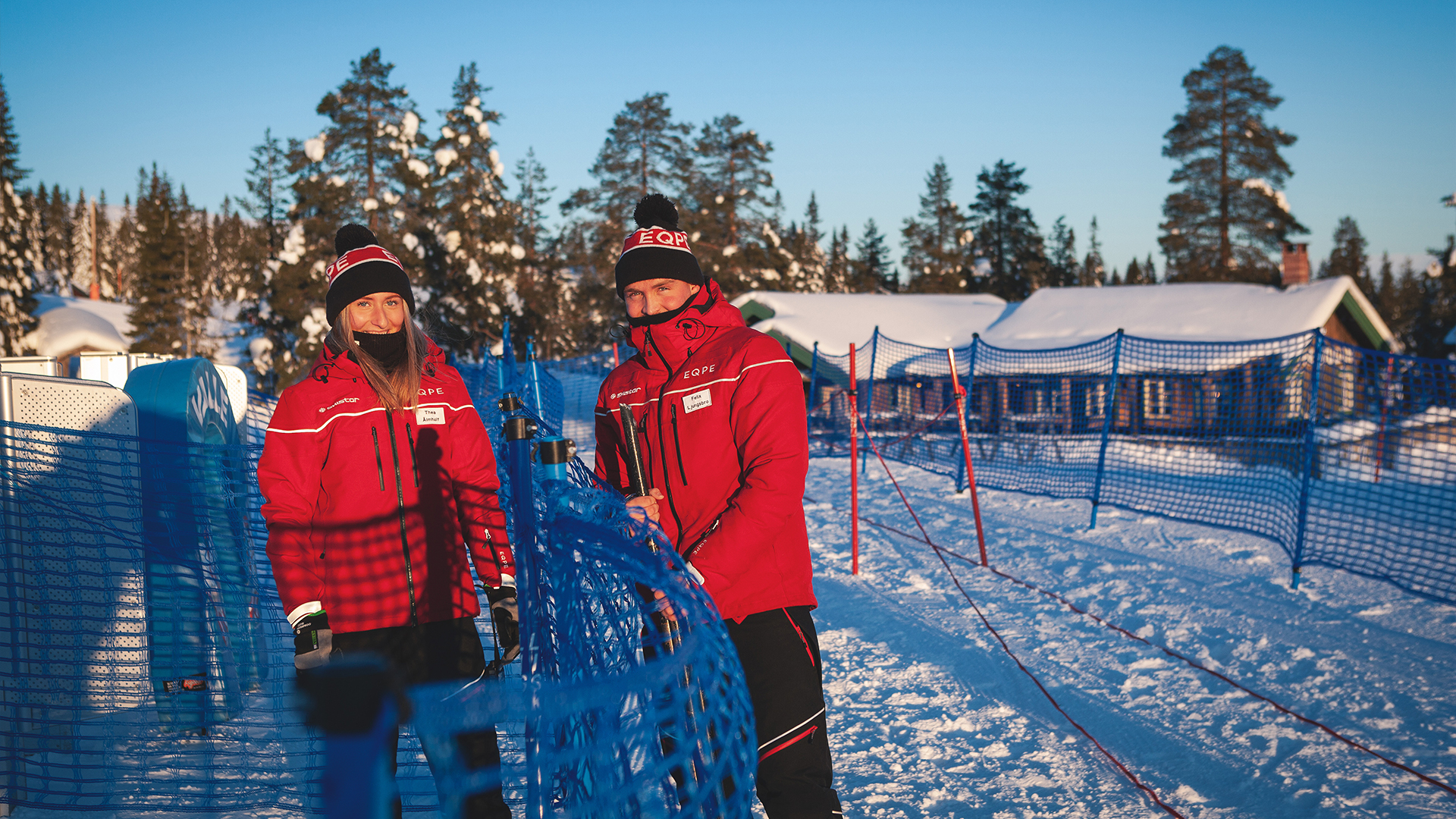 To personer i røde jakker med skihatter på, står ved et nett langs en skiløype i en snødekt vinterpark med hytter i bakgrunnen. Klar blå himmel og solskinn