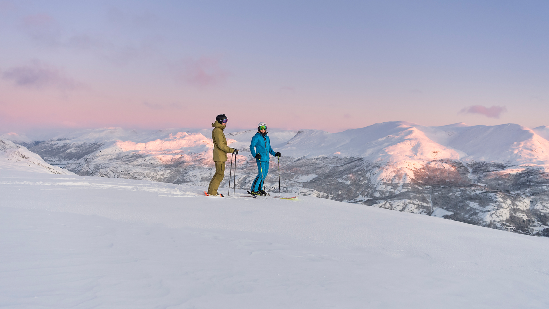 Två skidåkare står på en snötäckt bergstopp med en solnedgång i bakgrunden, fjällandskap i Norge