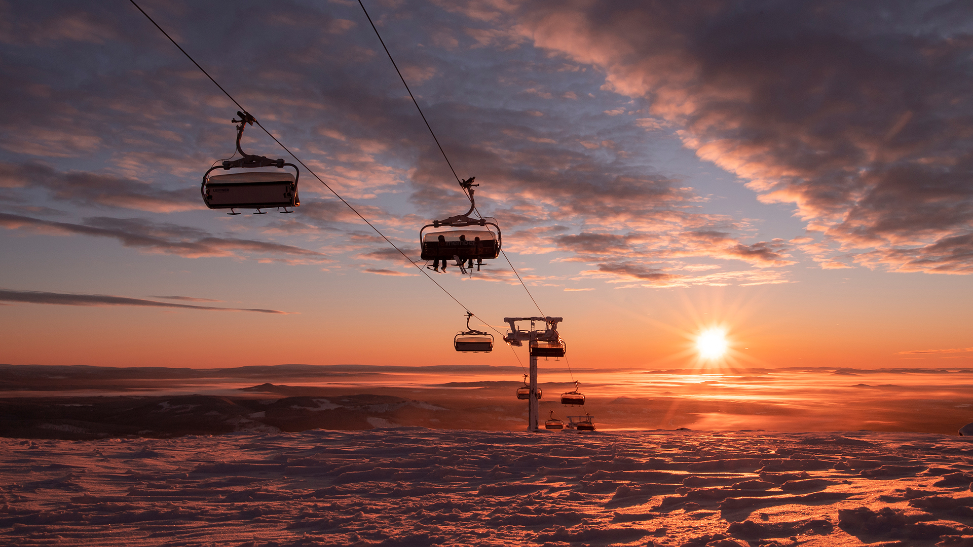Sessellift bei Sonnenuntergang über verschneiter Winterlandschaft, Wolkenhimmel und Sonnenstrahlen im Hintergrund