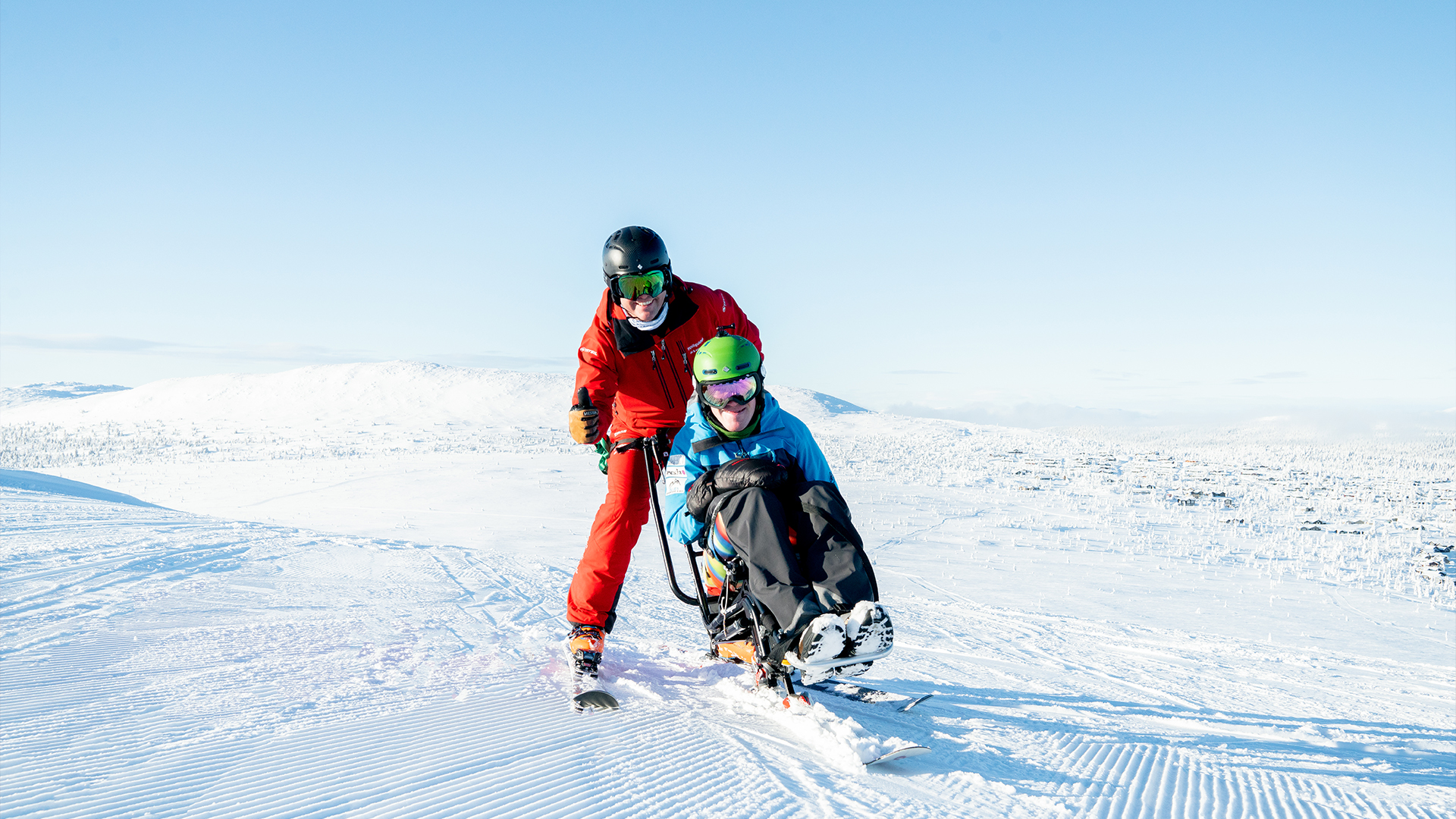 Två personer åker sitski på en snötäckt sluttning under en klarblå himmel. Den bakre personen i röd skidutrustning hjälper den främre personen i blå jacka och grön hjälm