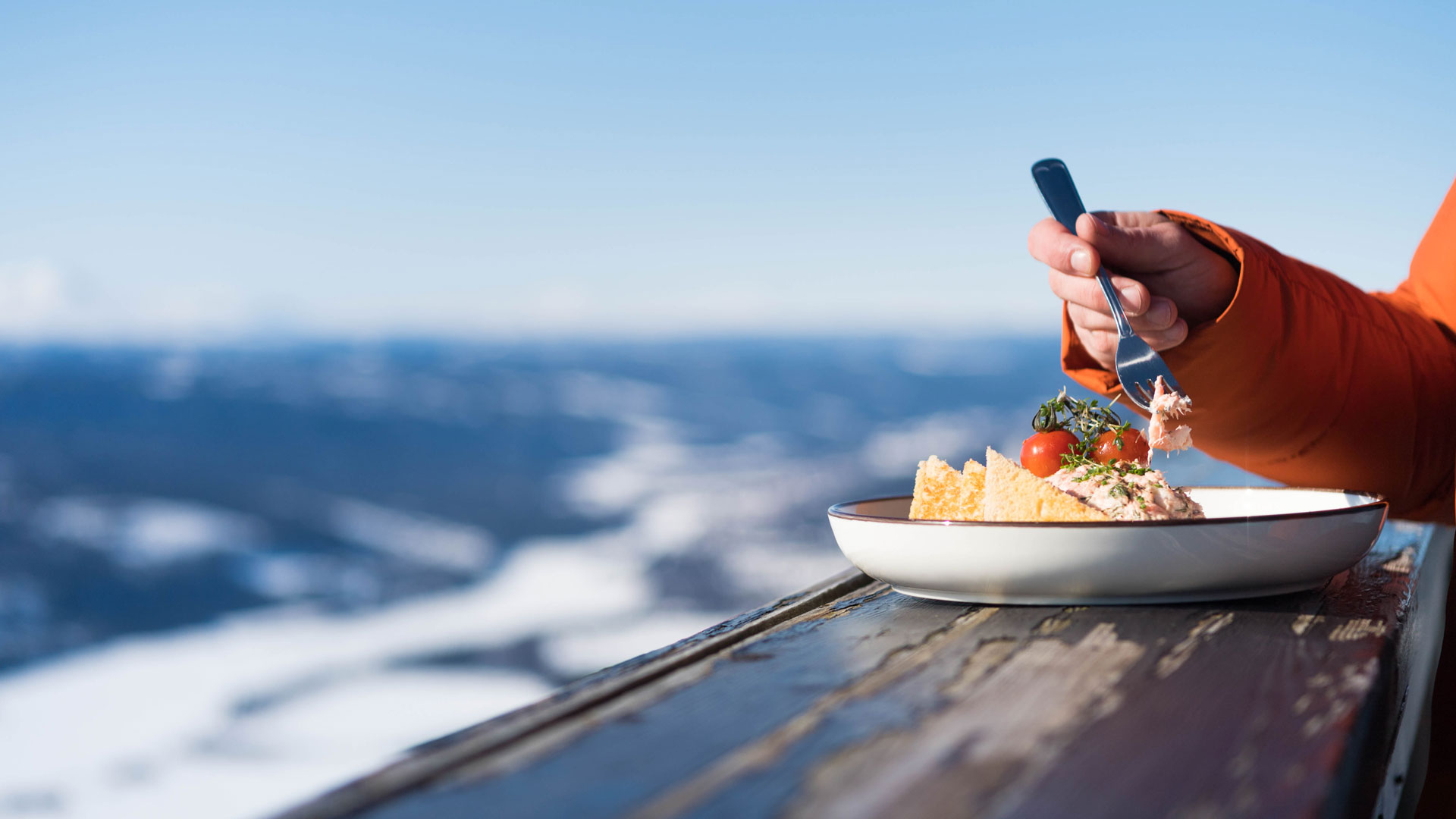 Person, der spiser en ret med tomater og sprøde chips på en tallerken, med sneklædte bjerge i baggrunden