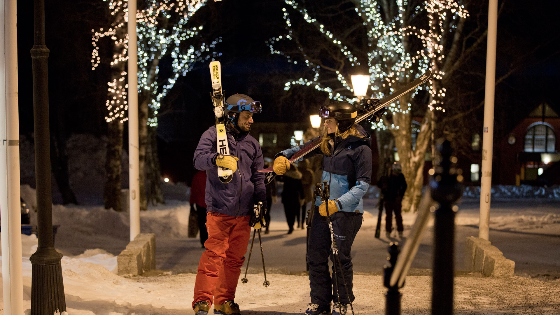 Två personer klädda i skidkläder bär skidor på axeln i en vintermiljö med snö och julbelysning på kvällen