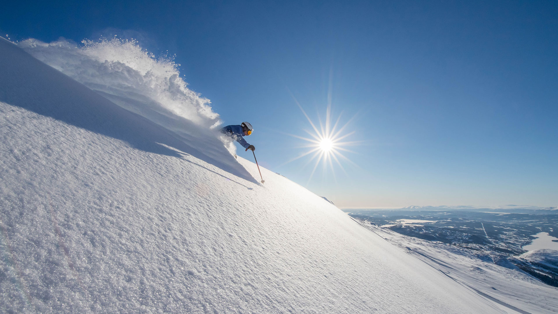 Skidåkare åker nerför snötäckt sluttning på solig dag i fjällen, med klarblå himmel och bländande solsken i bakgrunden