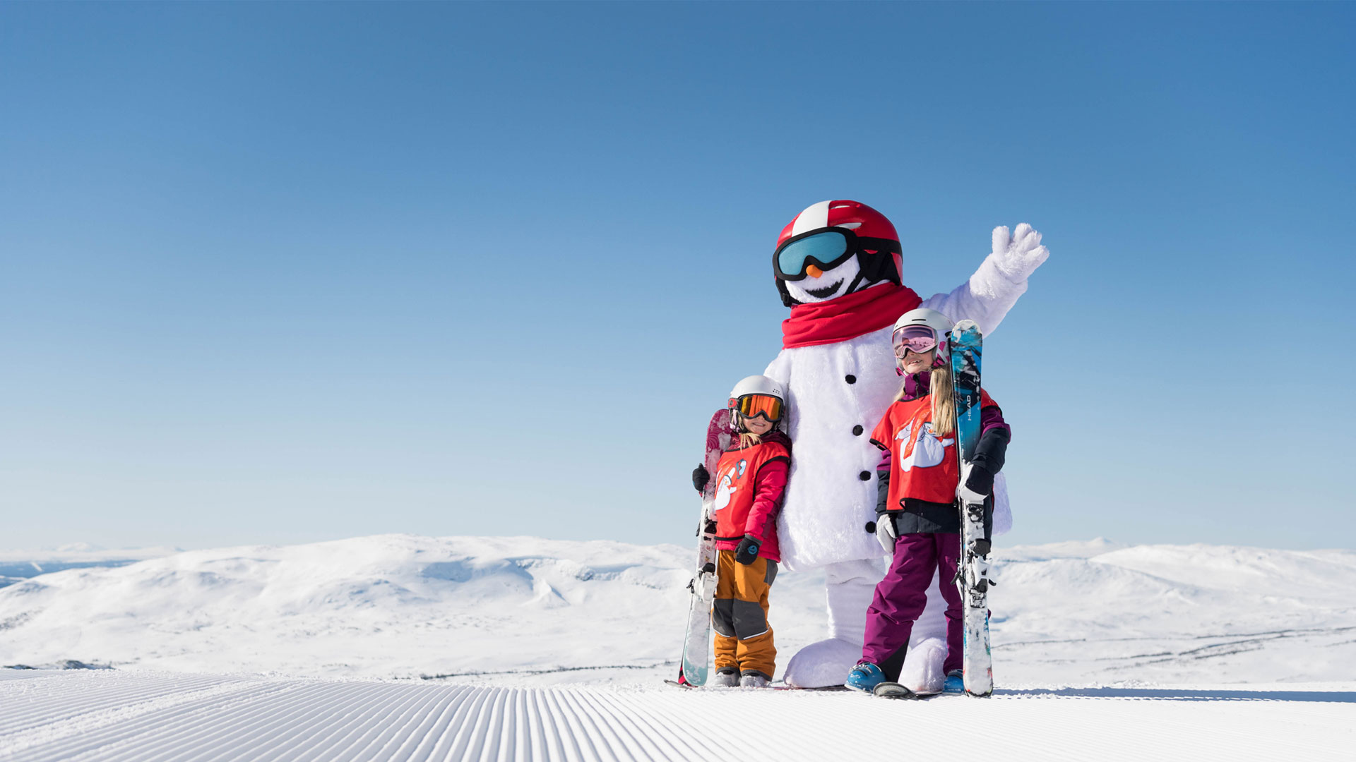 Snögubben Valle och barn i skidkläder poserar med skidor på en snötäckt bergstopp under en klarblå himmel