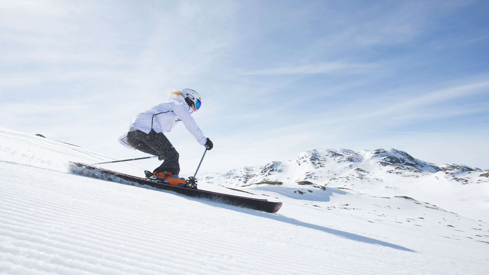 Person åker skidor nerför en snötäckt backe under en klarblå himmel med berg i bakgrunden