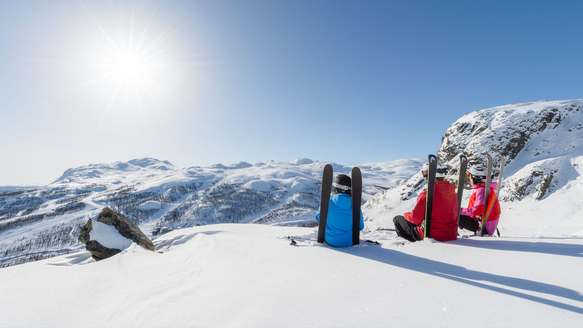 Två skidåkare sitter i snön med skidor restade framför dem, med en storslagen utsikt över snötäckta berg under solig himmel i bakgrunden