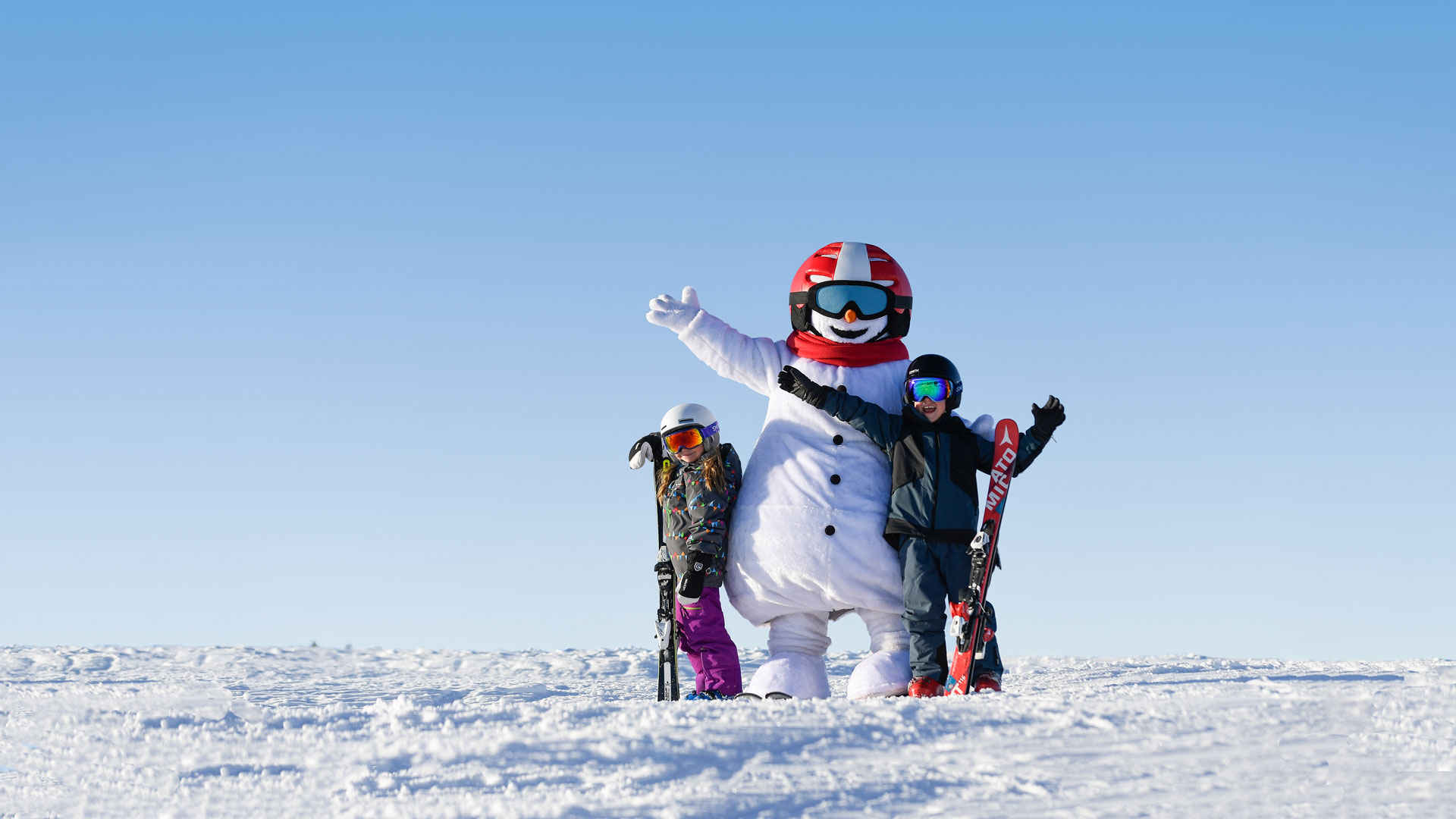 Snögubben Valle i skidutrustning poserar med två barn på en skidbacke under klar himmel