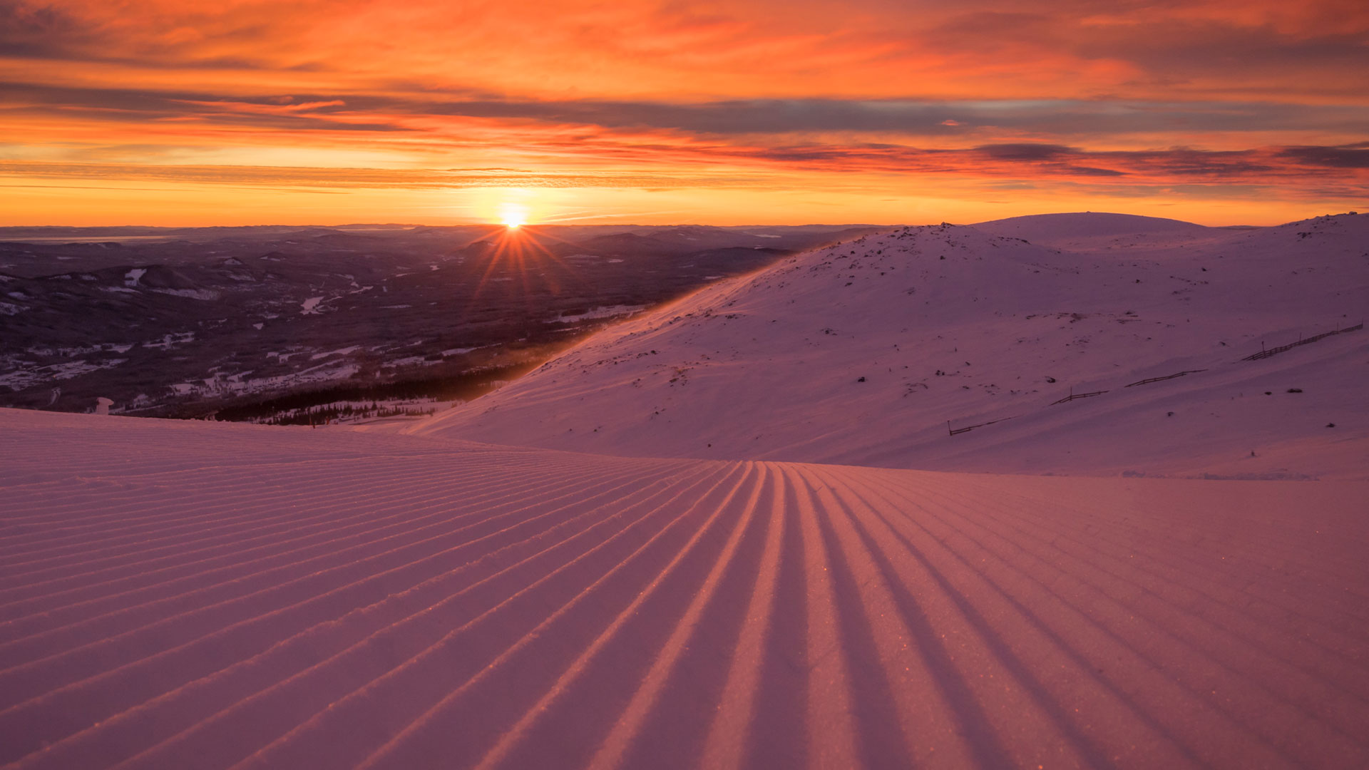 Solnedgang over snedækket bjerglandskab med lyserød og orange himmel. Solen går ned bag bjergtoppene, hvilket skaber et dramatisk lys på den præparerede sne i forgrunden