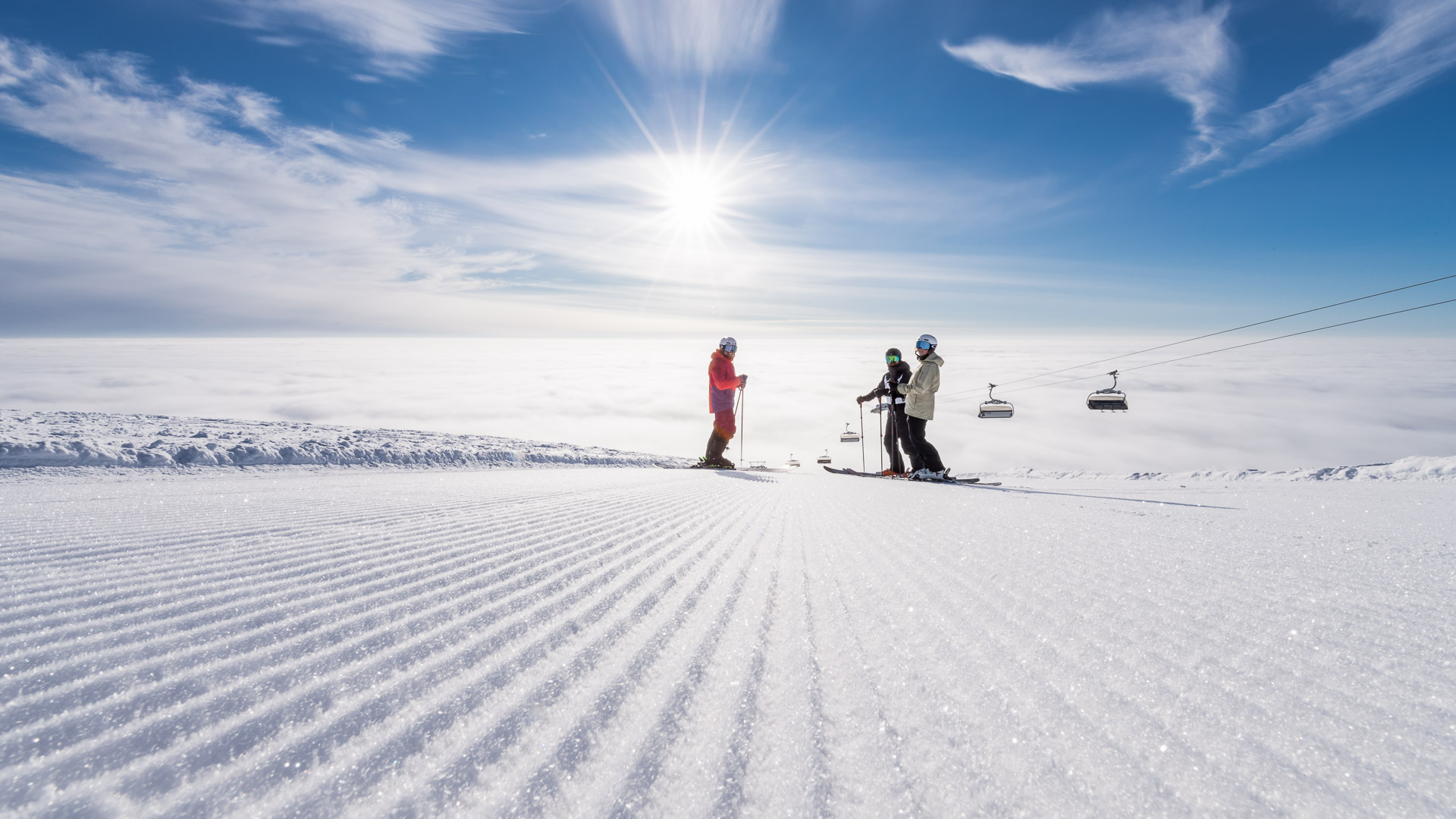 Tre skiløbere står på pisten under en solrig dag med skyer og blå himmel, ved siden af en stolelift