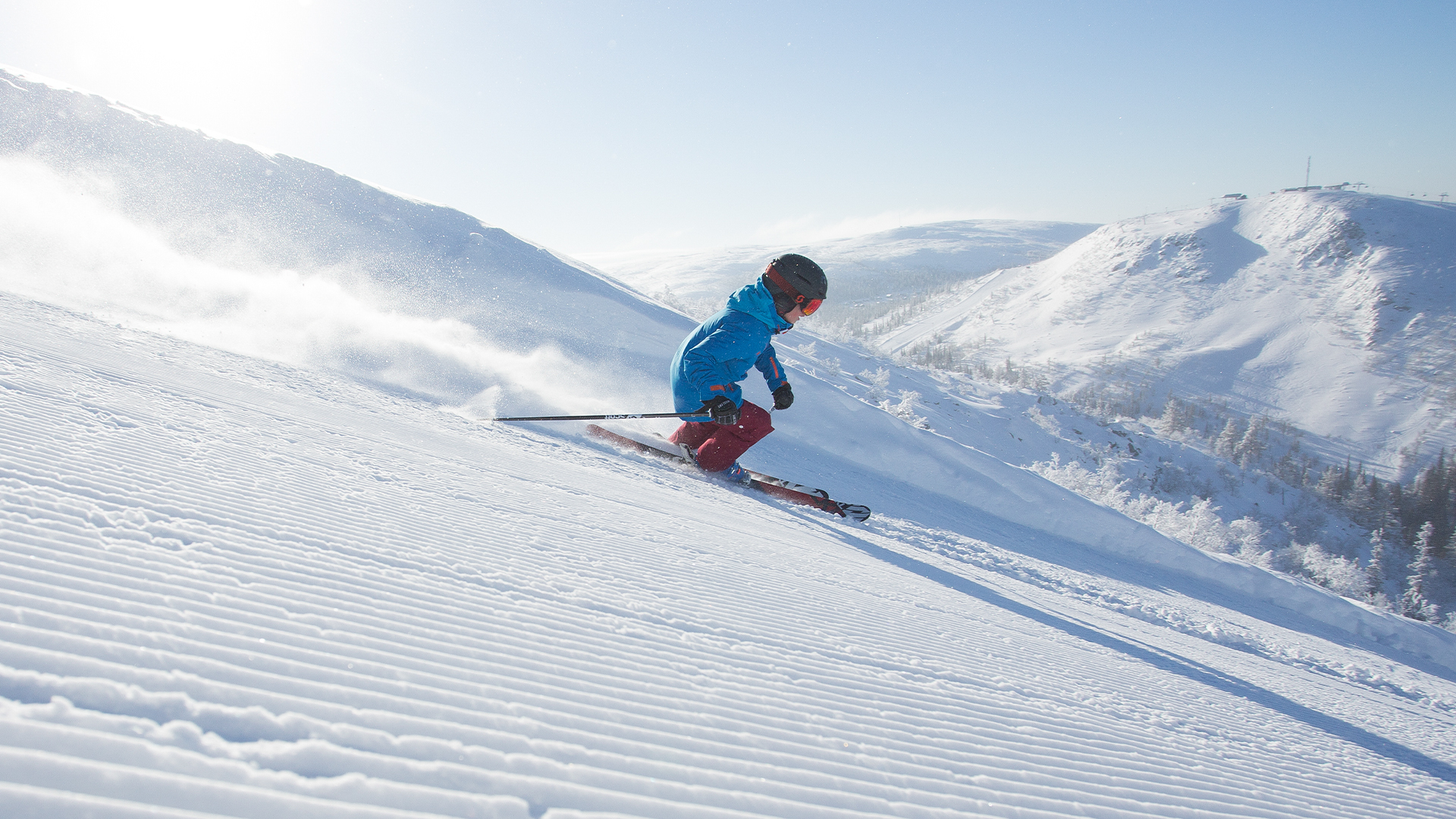 Person åker skidor nerför en snötäckt skidbacke i ett soligt bergslandskap