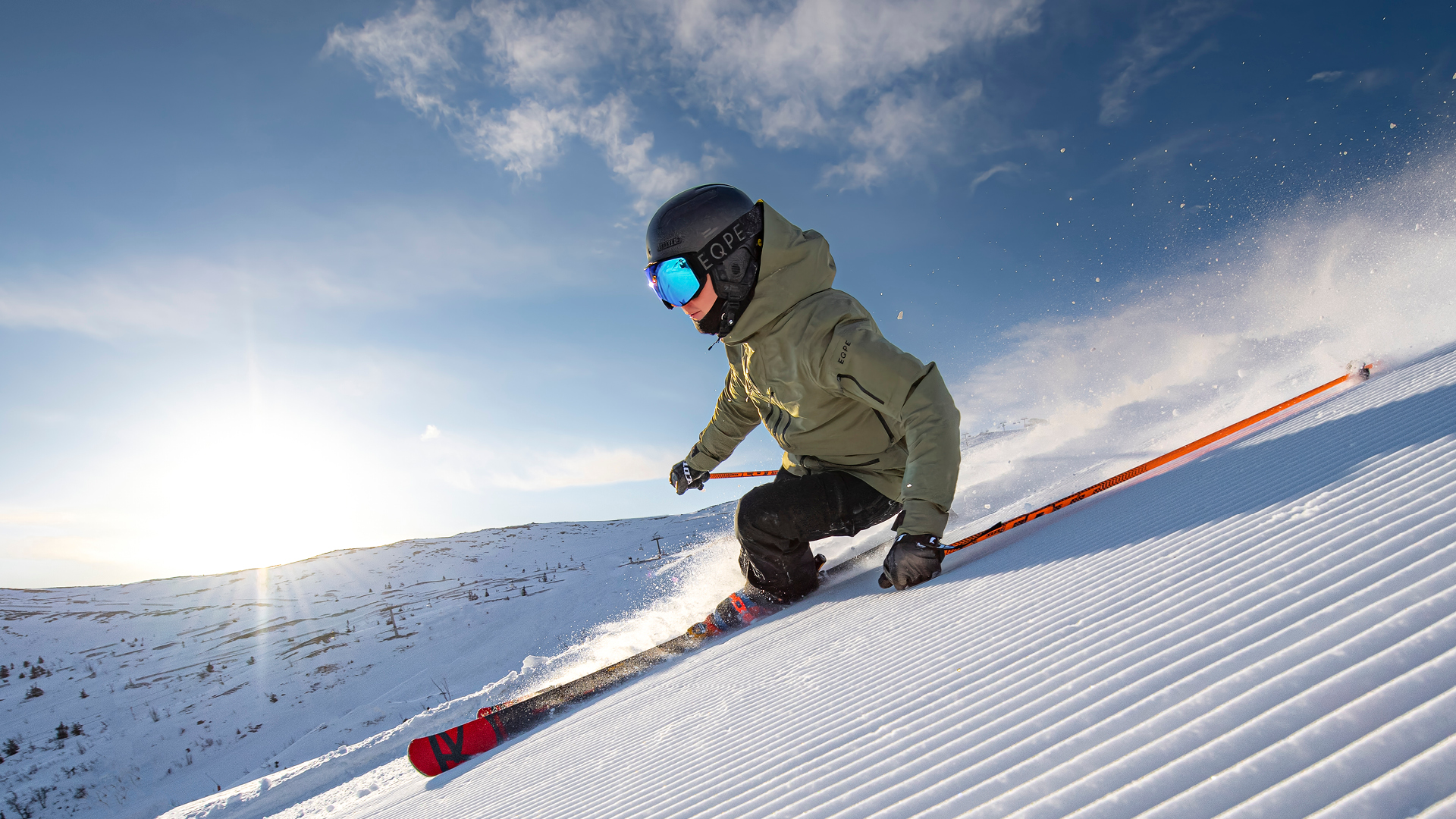 Man kjører på ski i høy fart nedover en nypreparert bakke under en klarblå himmel