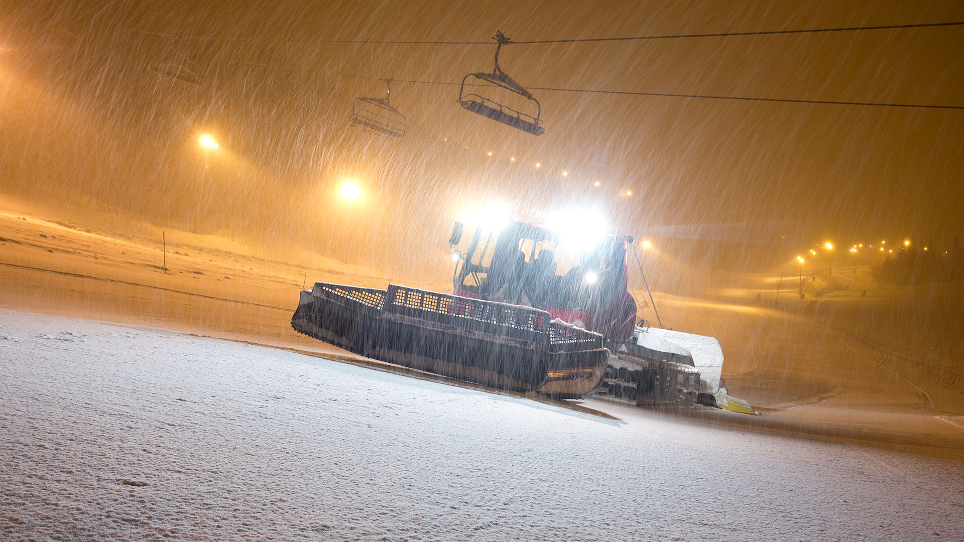 Schneepflug im Einsatz auf einer Skipiste während nächtlichem Schneefall, kräftige Lampen erhellen die Umgebung