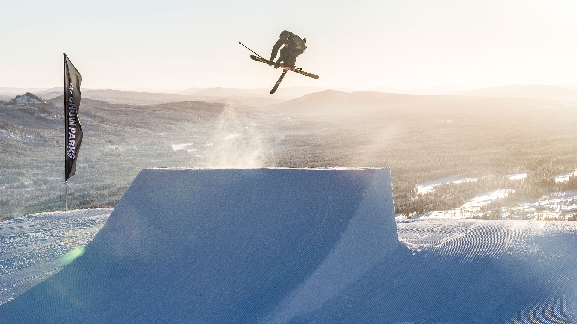 Skidåkare utför en vågad hopptrick i en solig snöpark med vy över snötäckta fjäll i bakgrunden