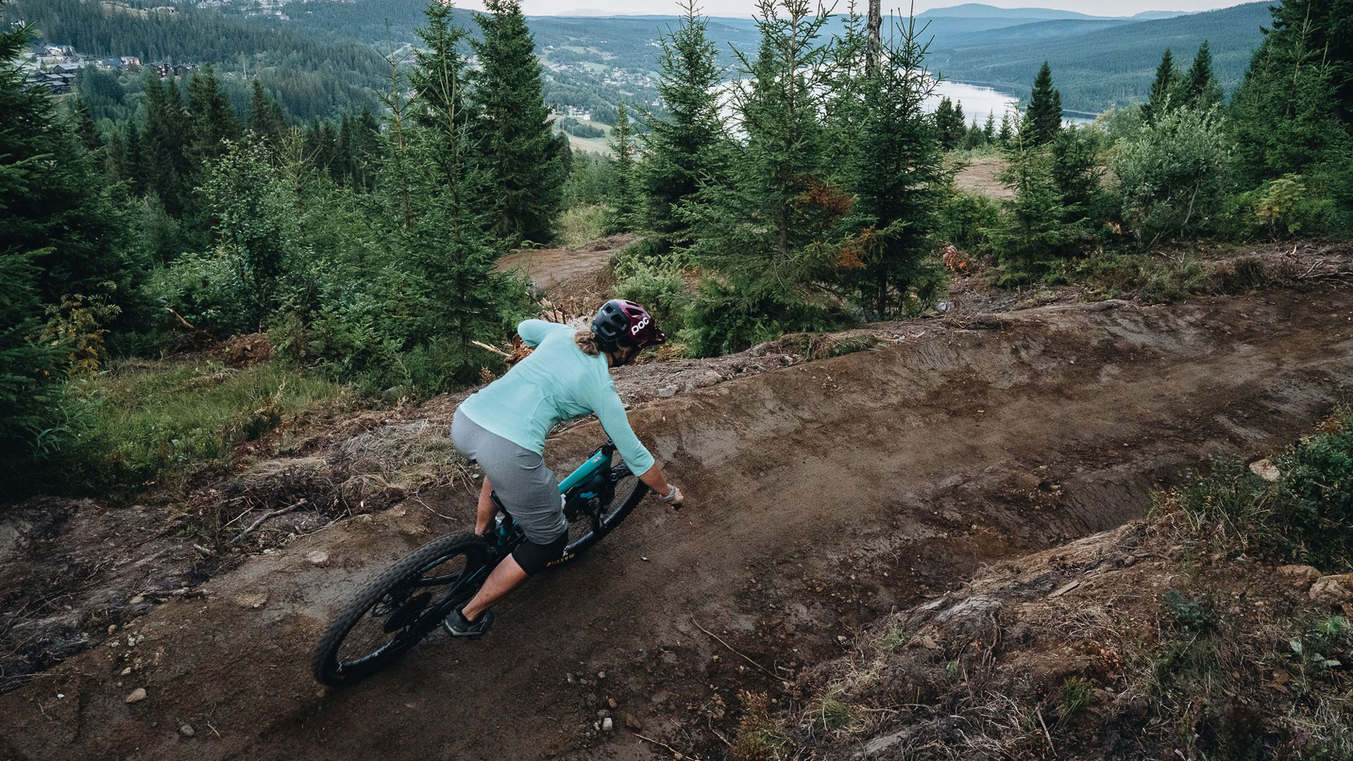 Mountainbikeåkare på skogsstig i fjällmiljö, omgiven av grönskande träd och berg i bakgrunden