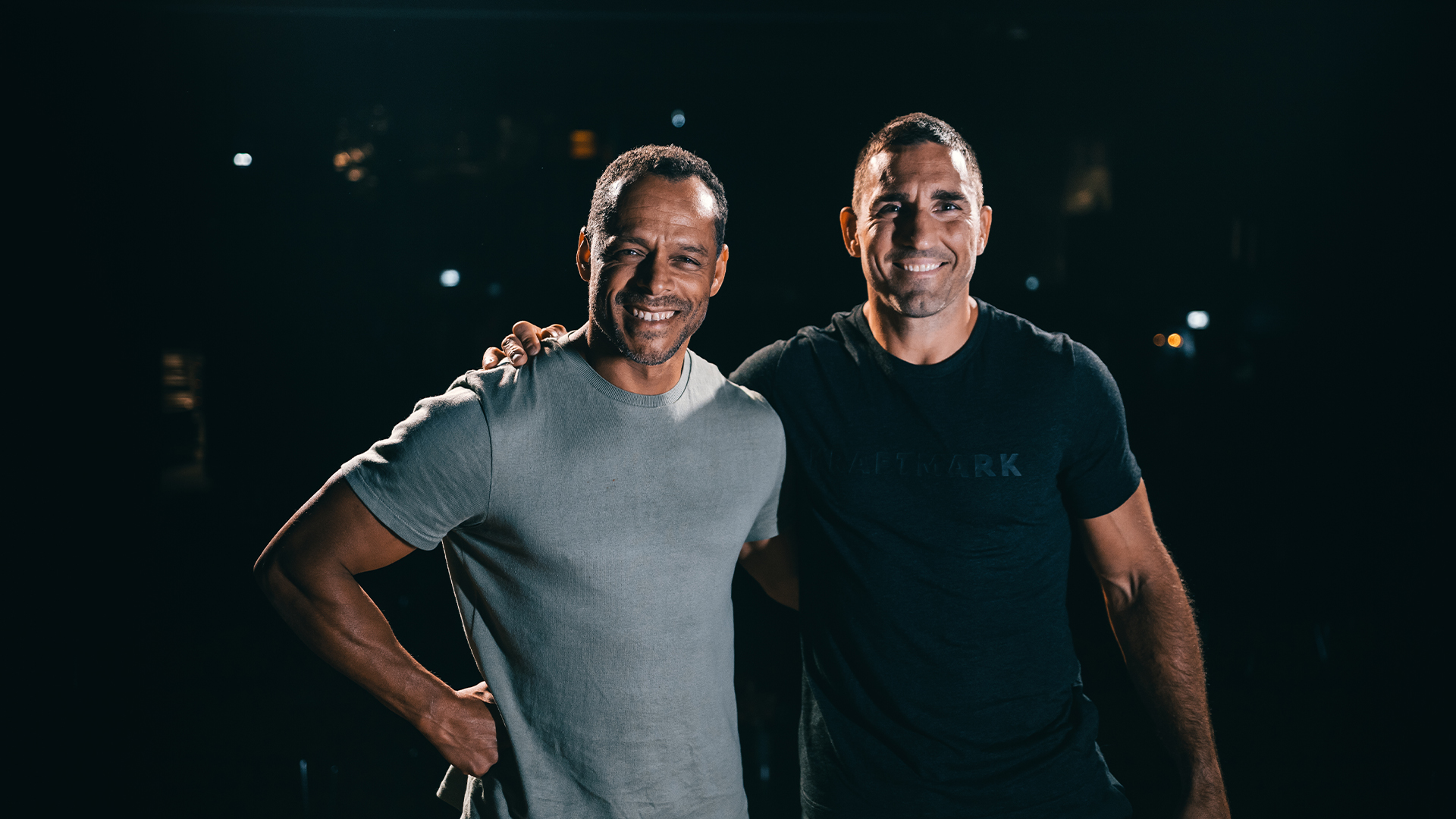 To smilende mænd i træningstøj står sammen på en mørk baggrund