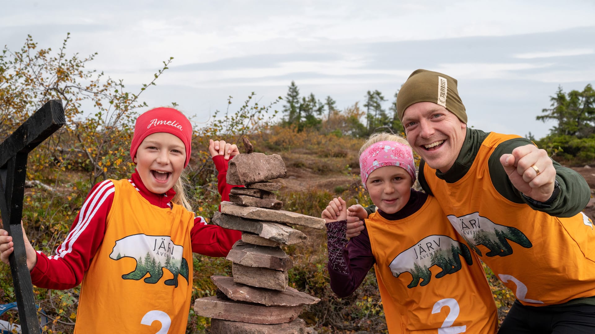 Tre personer deltar i utomhusaktiviteten Järväventyr, iklädda orange västar, i en naturskön miljö. De ler och poserar vid en stapel stenar