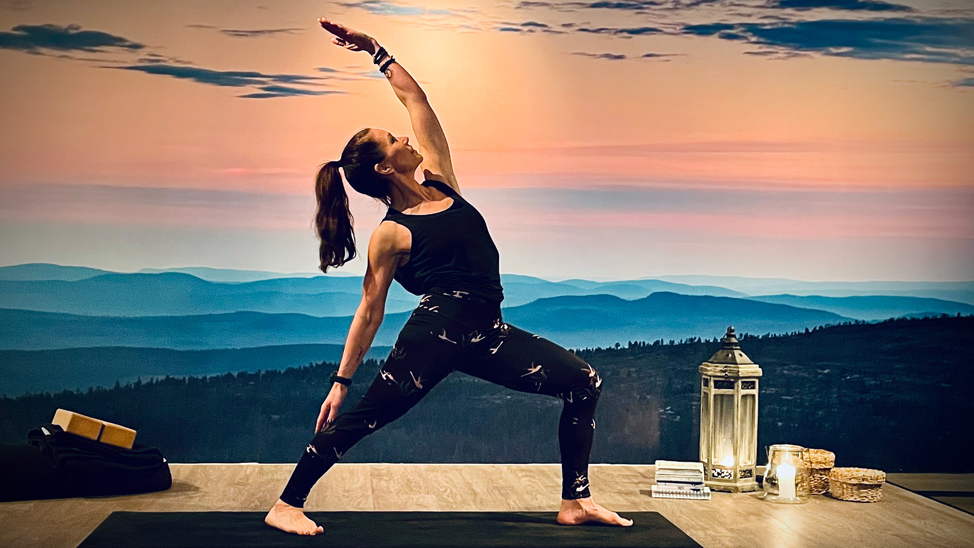 Kvinna utför yoga i krigare II-position i solnedgång med blå berg i bakgrunden, med ljus och lantlig inredning på golvet