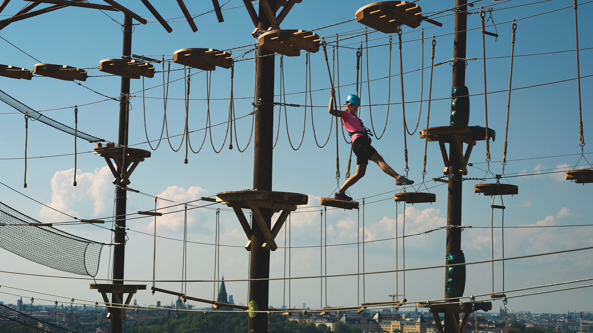 Person klädd i hjälm och sele som balanserar på en hög äventyrsbana med rep och plattformar, mot en blå himmel