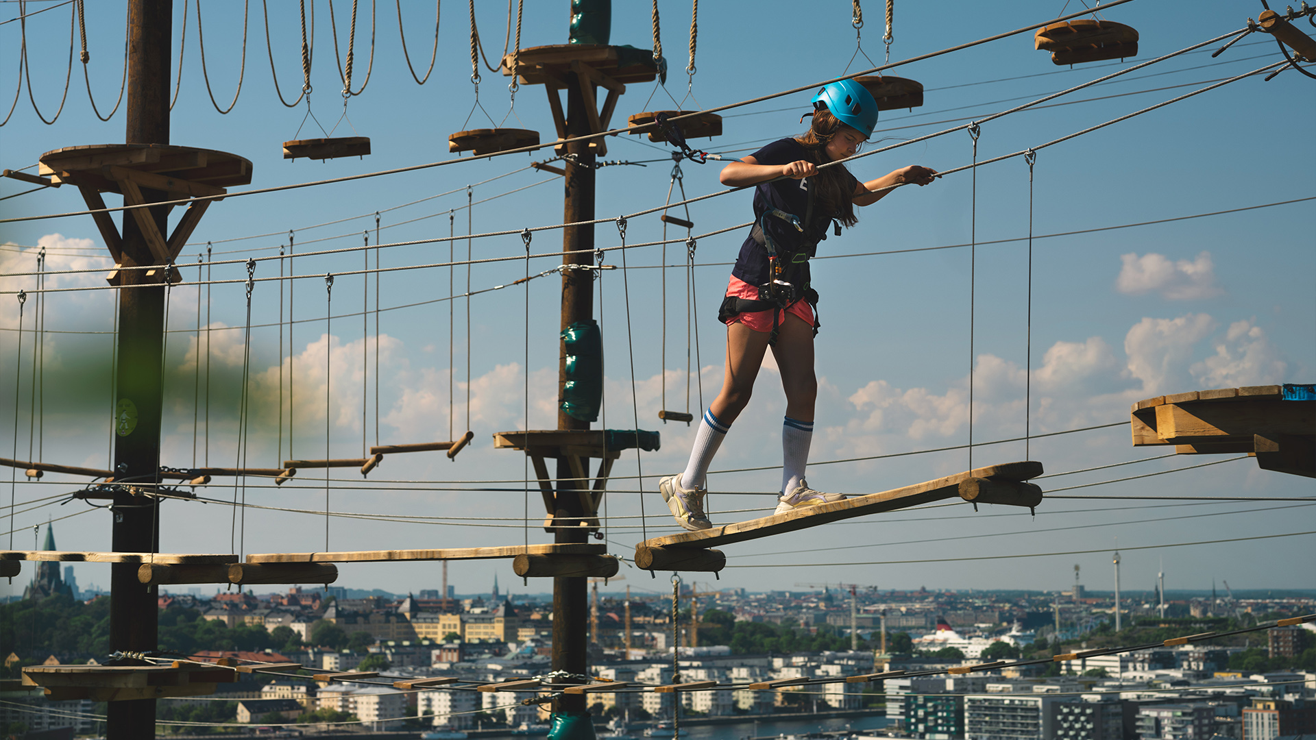 Kvinna iförd blå hjälm går en höghöjdsbana ovanför stadshorisonten på en solig dag