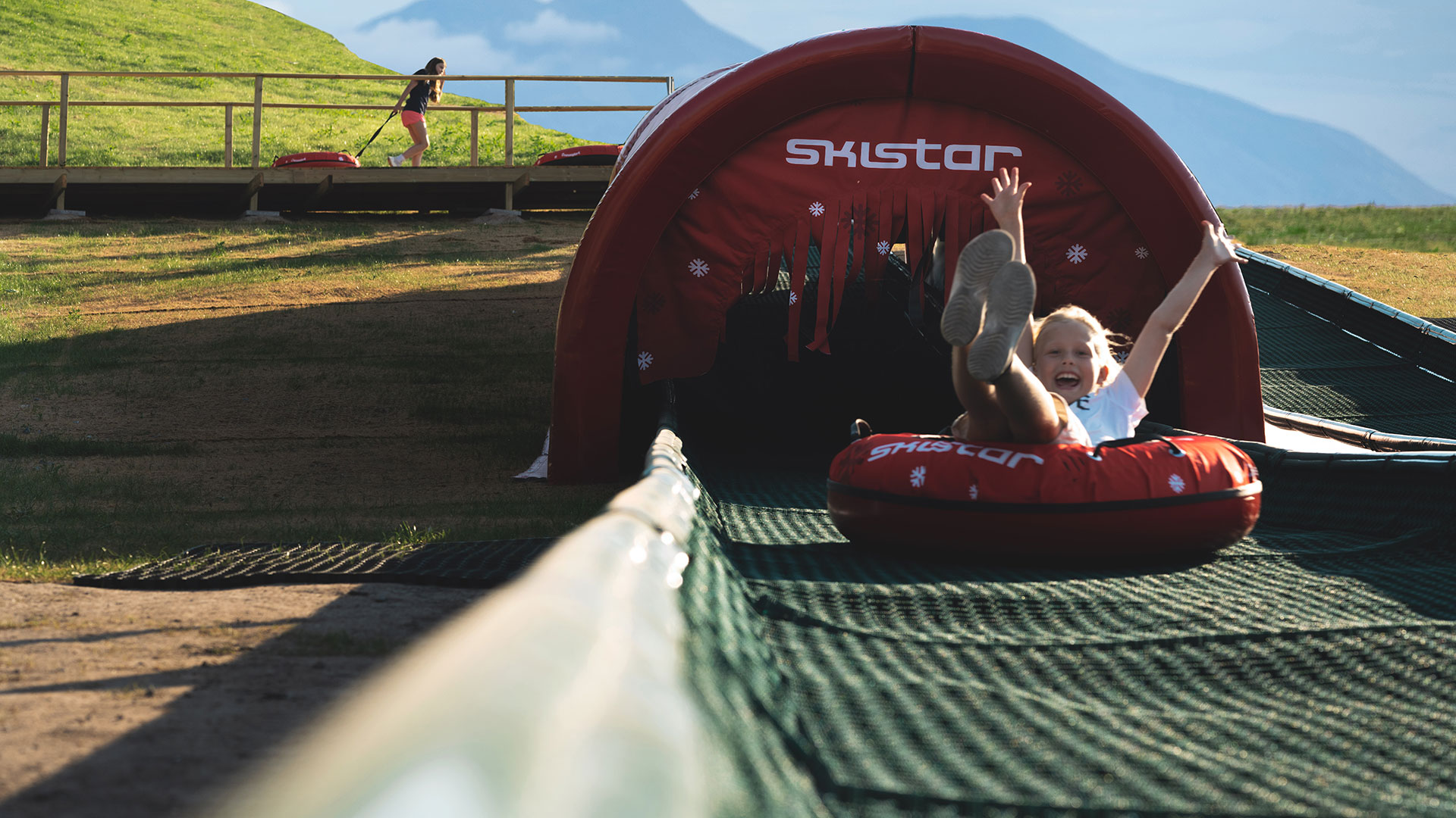 Person i en rød skliering glir nedover en sommerrodelbane under en solrik dag, omgitt av grønne felt og en blå himmel