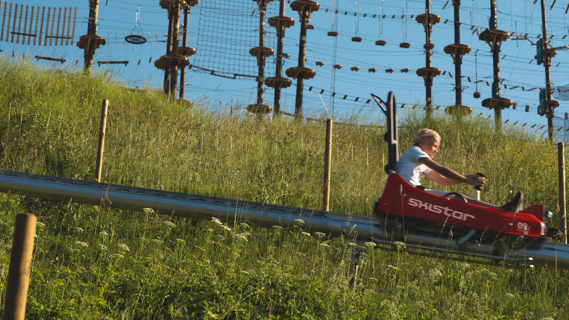 Person åker sommarrodel nerför en gräsbevuxen sluttning på Skistar med höghöjdsbana i bakgrunden