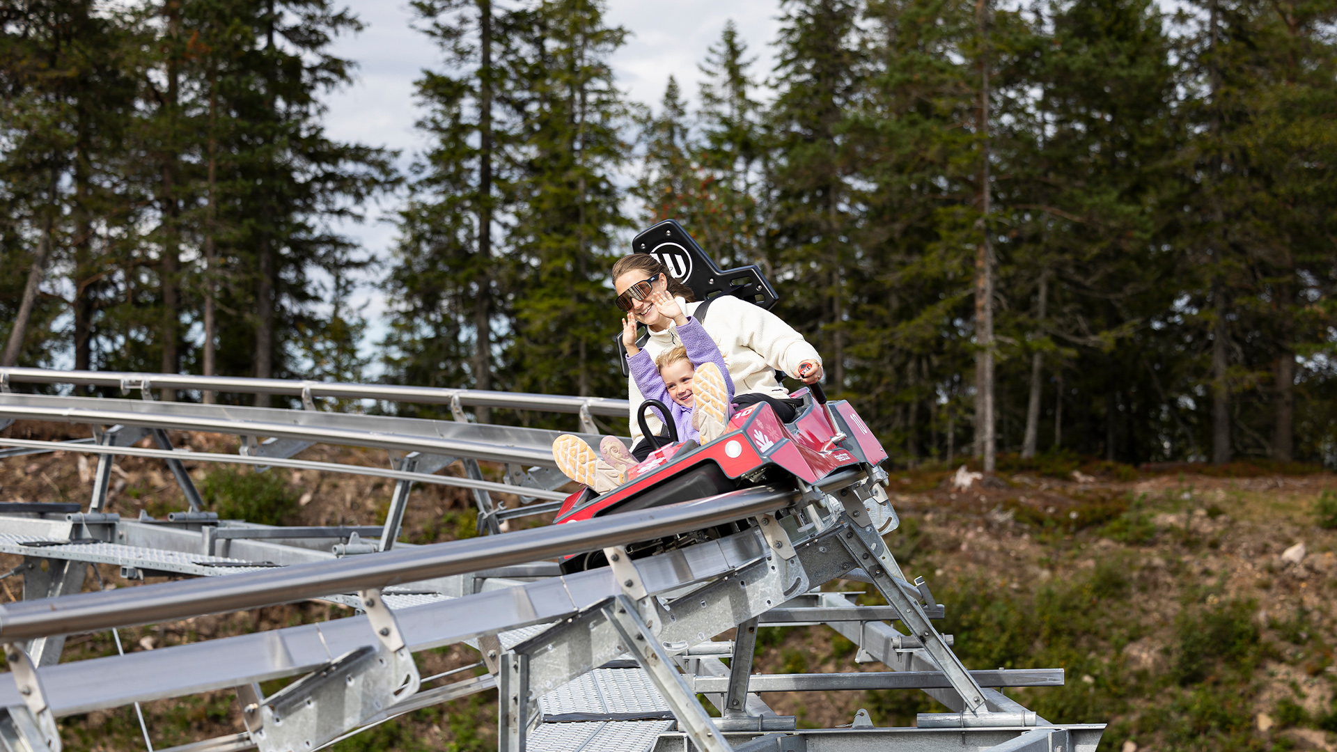 Personer åker Mountain Coaster, en typ av rodel, på en räls i Lindvallen, Sälen. Det är en mamma och ett barn och de ser glada ut.