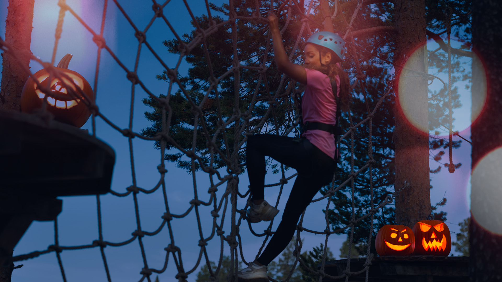 Barn klättrar i nät med hjälm i skymningen, omgiven av Halloween-lyktor i form av pumpor, i en skogsmiljö