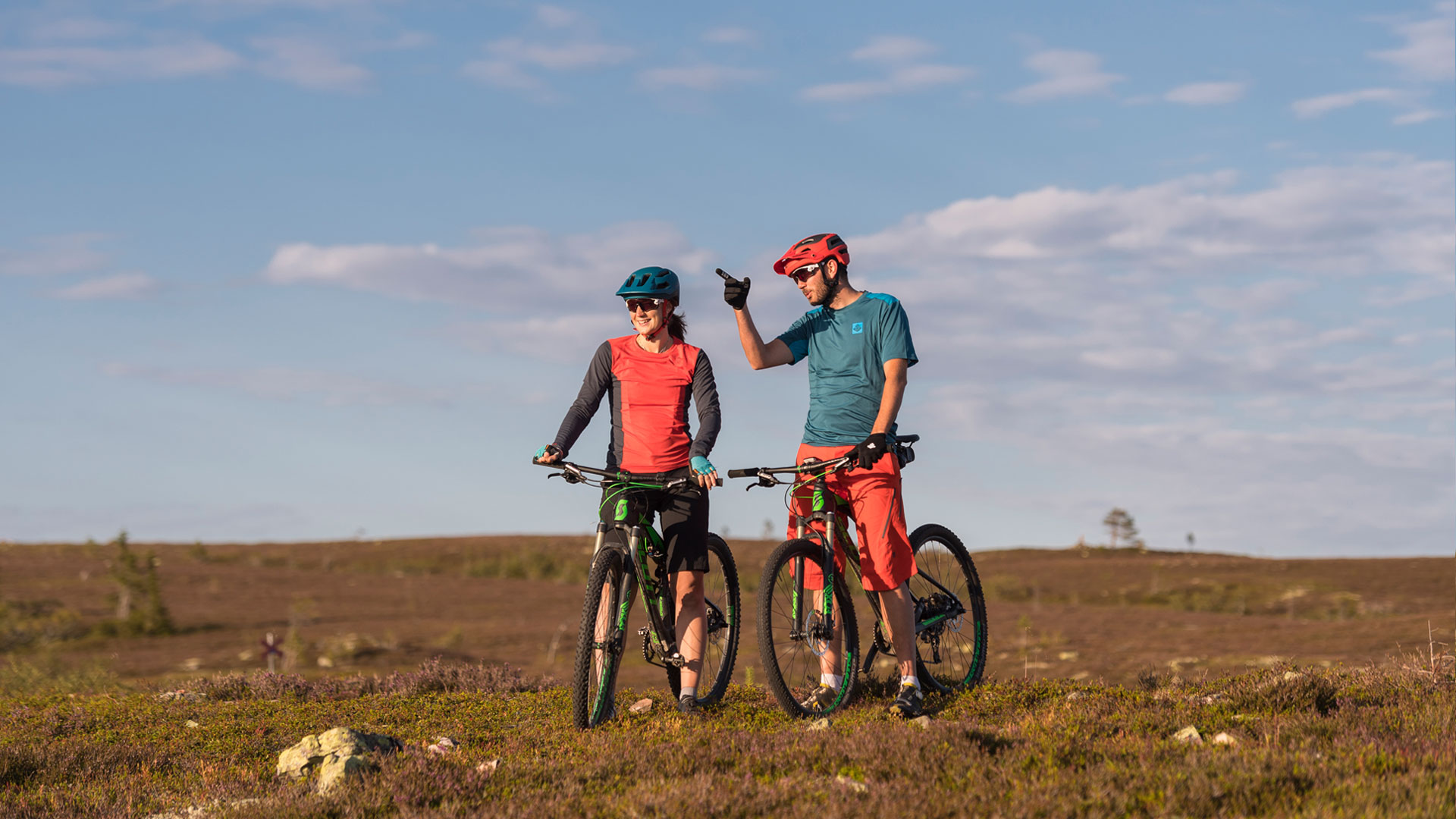 Två cyklister med hjälmar står med sina mountainbikes på en solbelyst hed och pratar under en klar himmel