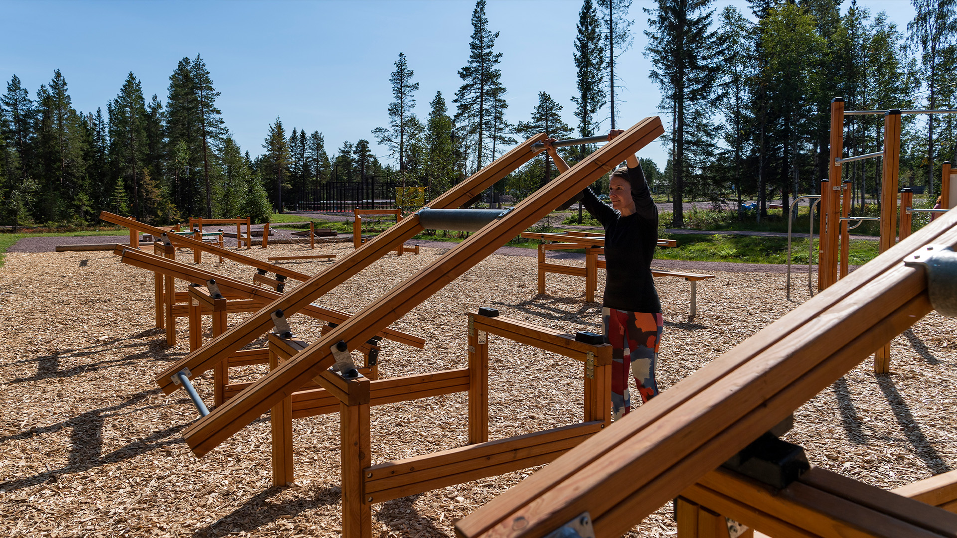 Utegym med träutrustning i en skogsmiljö, person tränar på station omgiven av träd i bakgrunden