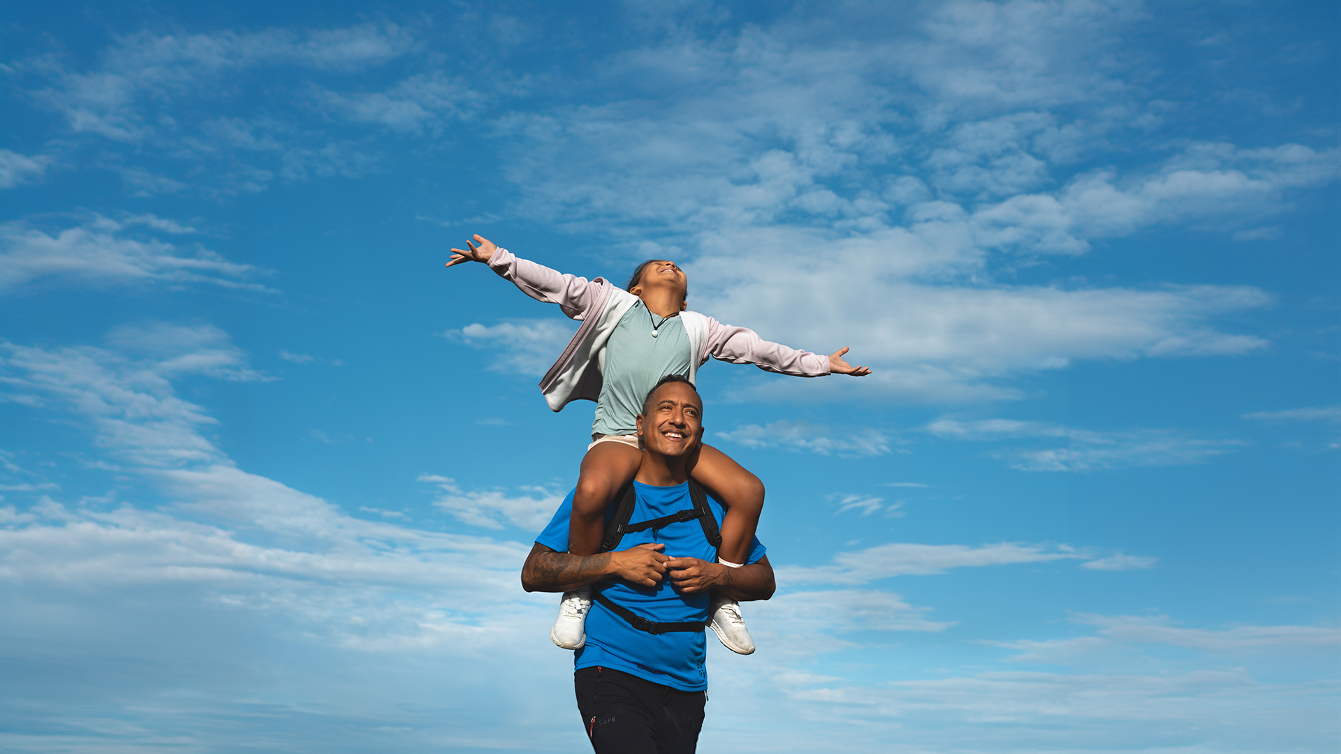 Vater und Tochter genießen das sonnige Wetter im Freien, die Tochter sitzt fröhlich auf den Schultern des Vaters vor einem klarblauen Himmel